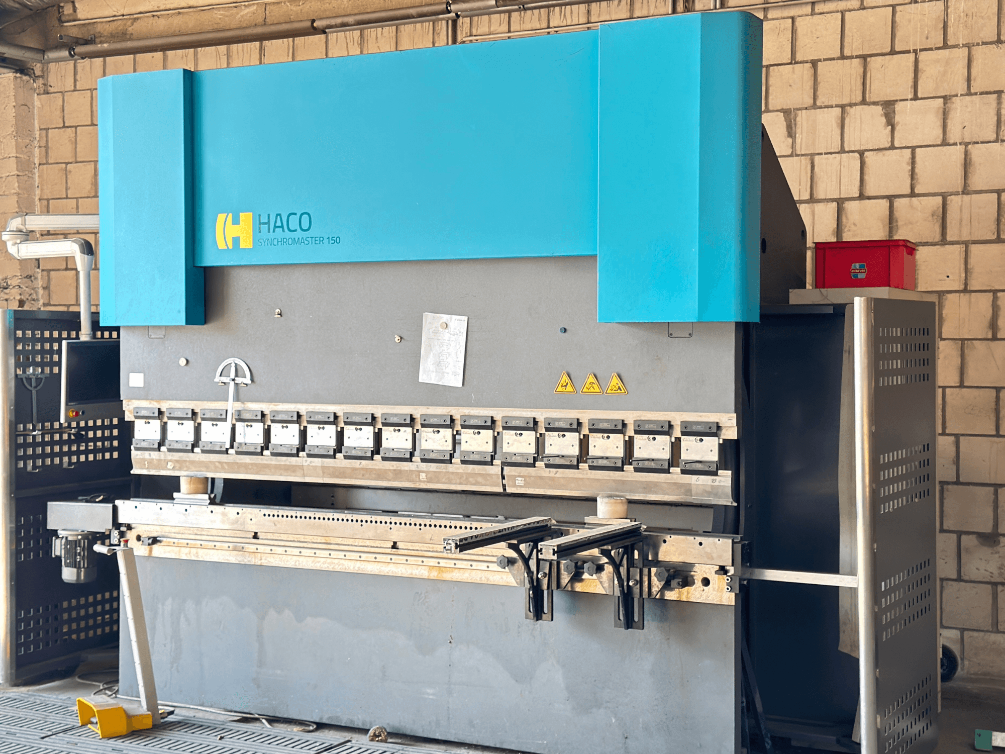 HACO SRM30150 abkant pres, önden görünüm; kontrol paneli ve sıkıştırma kalıplarını içerir, güvenlik uyarı etiketleri görünür.