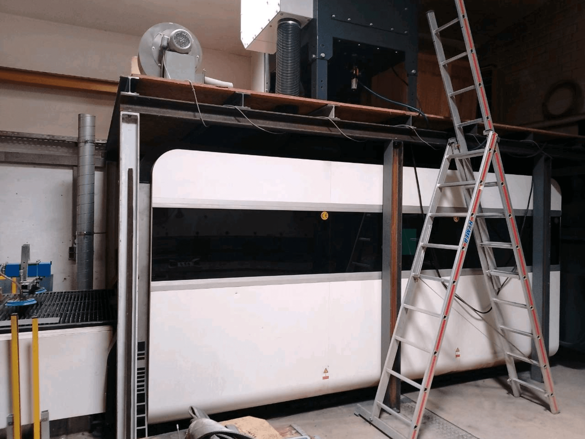 Bir atölyede bulunan GWEIKE LF3015GA fiber lazer kesim makinesi; ön görünüm, metal çerçeve ve erişim merdiveni gösterilmektedir.