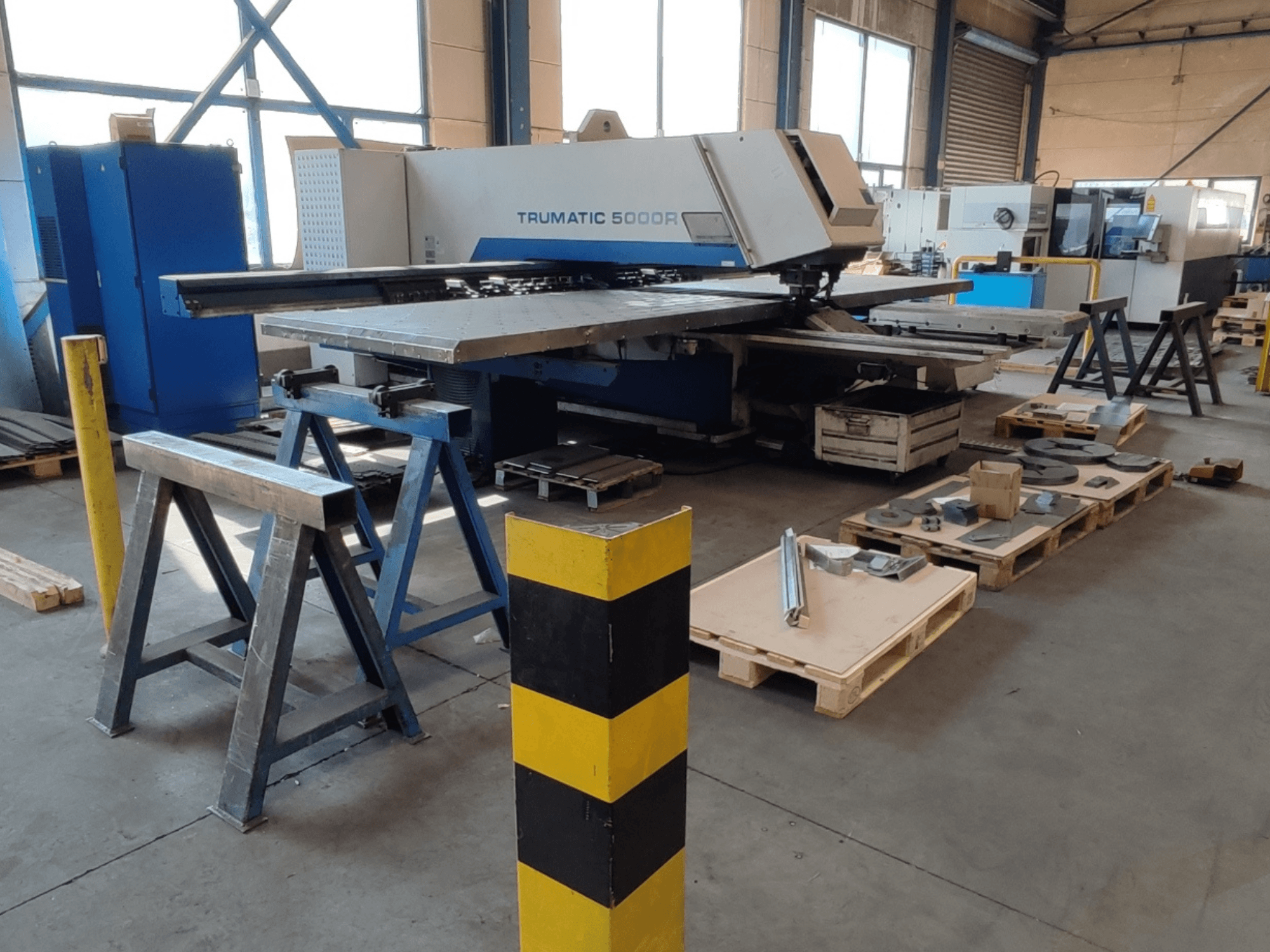 Bir atölyede bulunan TRUMPF Trumatic 5000 R CNC delme makinesi; yan görünümde, yakınında paletler üzerinde aletler ve malzemeler görülüyor.