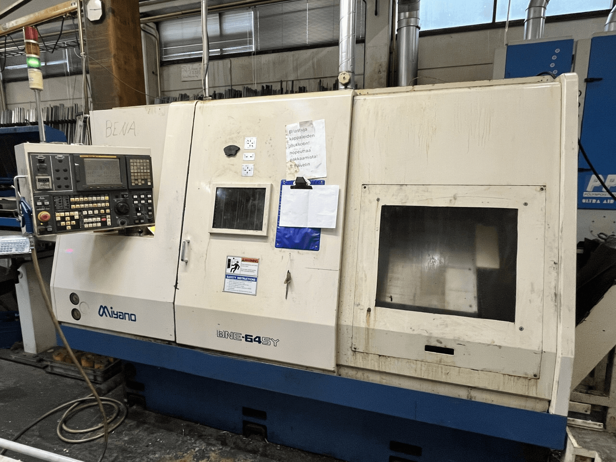 Miyano BNE-64SY CNC torna makinesi, önden görünüm, düğmeli kontrol paneli, LCD ve kabin üzerinde bakım etiketleri bulunmaktadır.