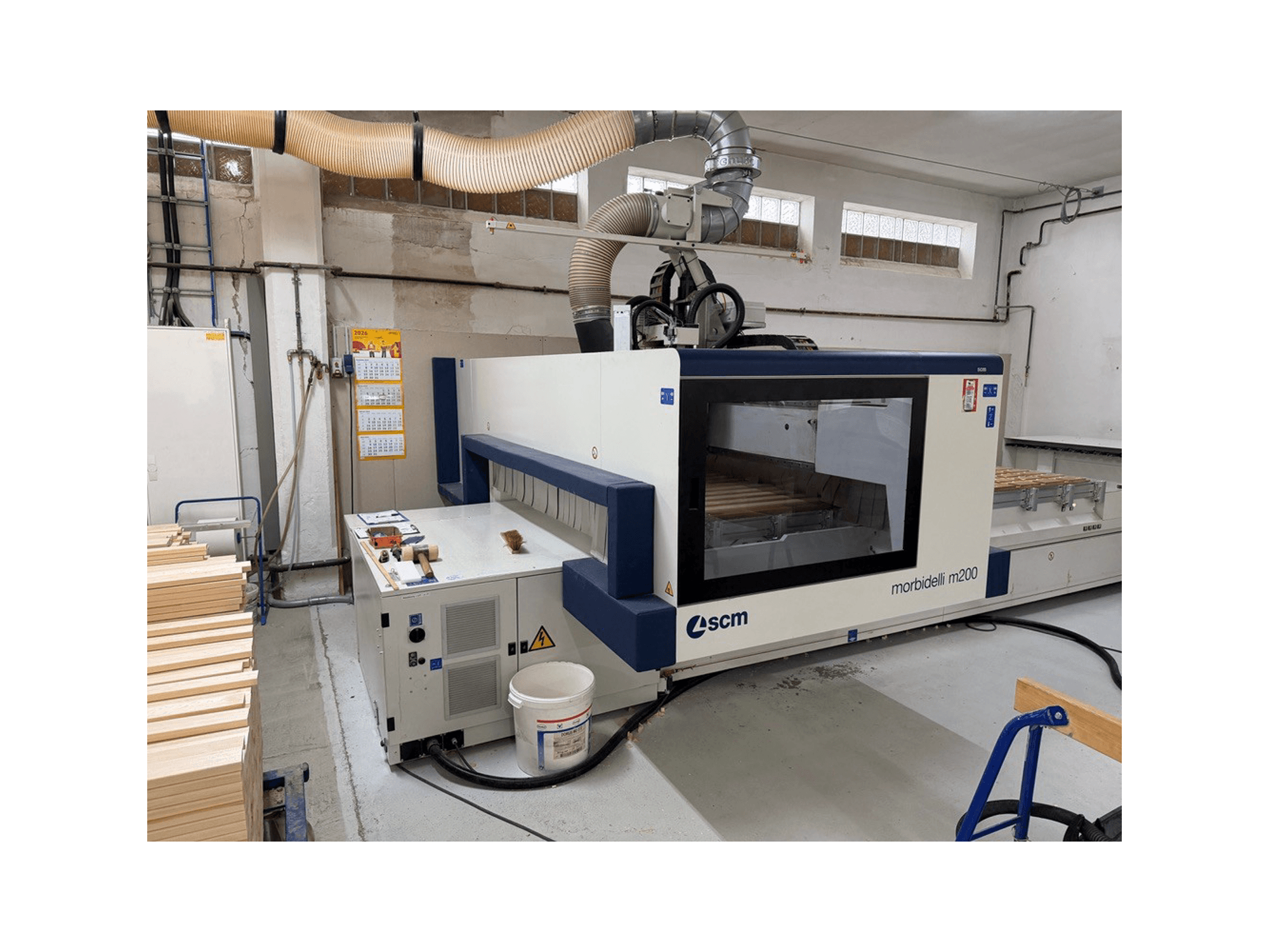Bir atölyede bulunan SCM Morbidelli M200F CNC işleme merkezi; masanın üzerinde ahşap parçalar ve üstünde havalandırma sistemi bulunan yan görünüm.