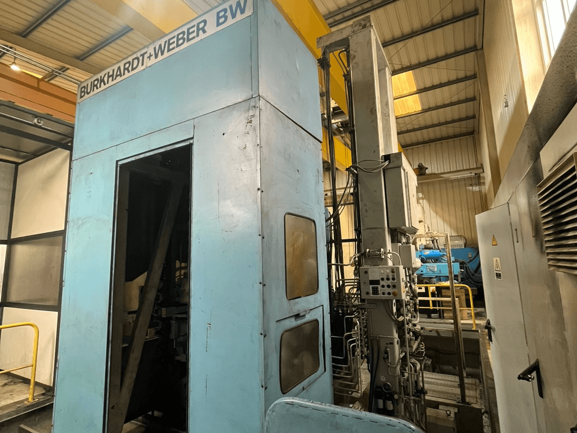 Burkhardt-Weber MC100 Yatay İşleme Merkezi; mavi dış yüzeyi ve üzerinde borular görülebilen kontrol panelini gösteren yan görünüm.