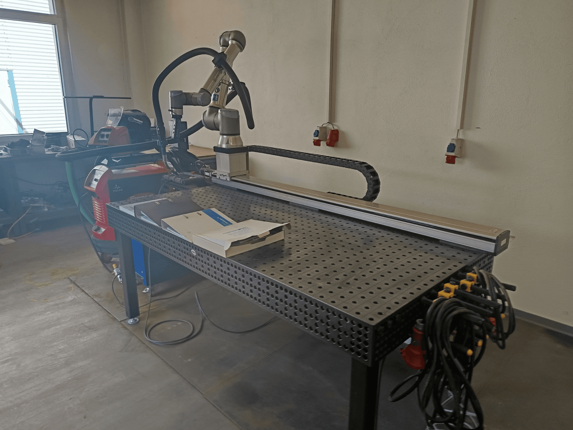 Yandan bakıldığında, bir robot kolu, çalışma alanı ve güç bağlantılarını içeren FRONIUS TransSteel 4000 Darbeli Kaynak Hücresi.