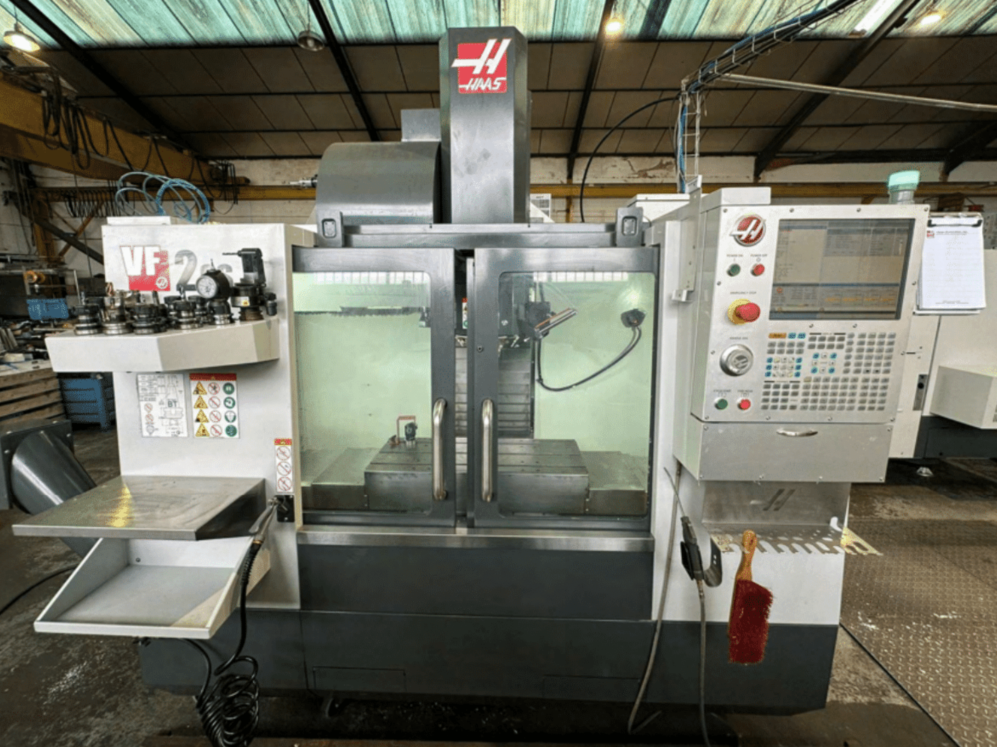 Haas VF2 CNC makinesinin önden görünümü, kontrol panelini, takım tutucuları ve güvenlik etiketleri ve bir fırça ile çalışma alanını gösterir.