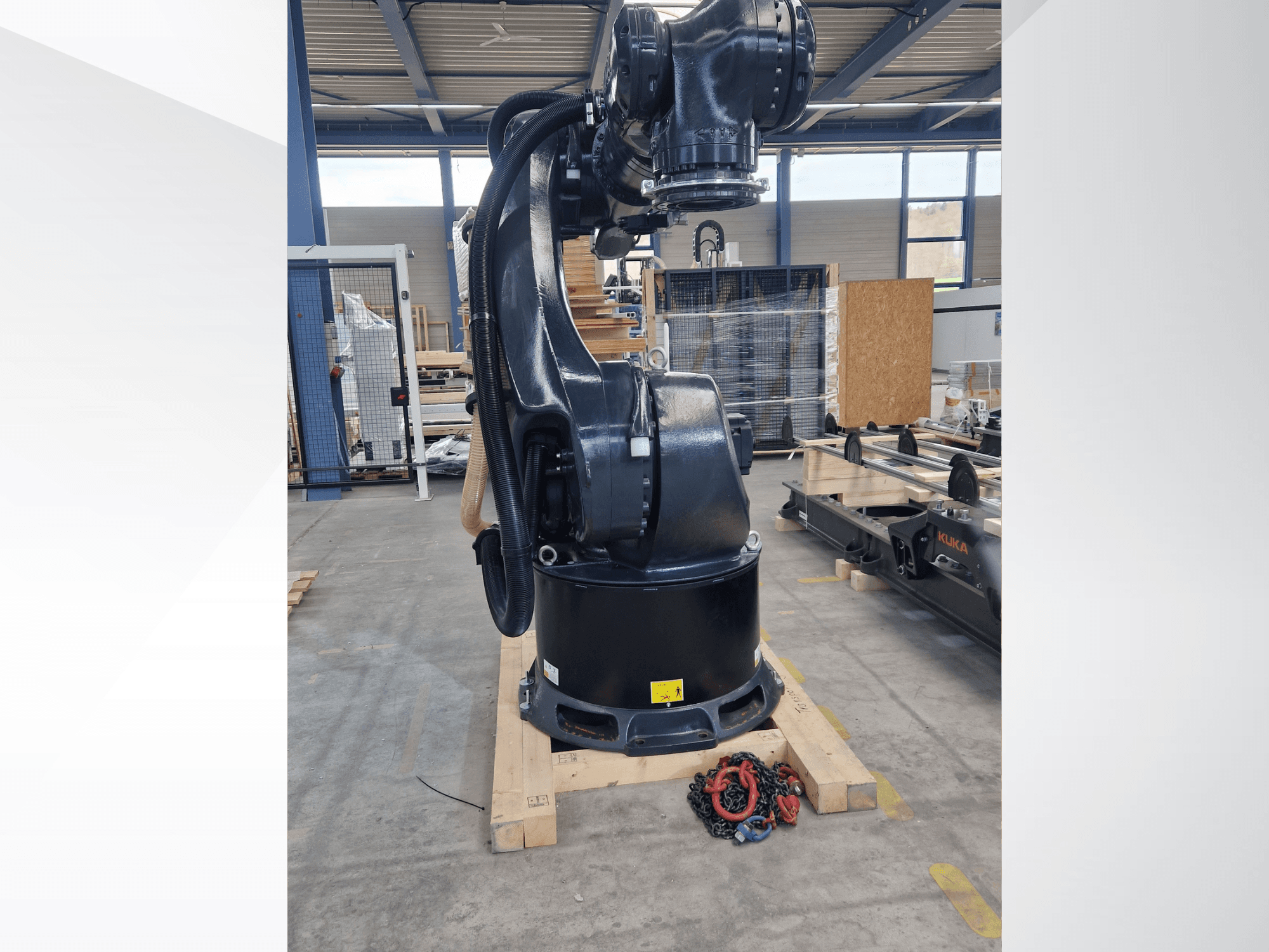 Bir depoda bulunan KUKA KR 340 R3330 robot kolu; tabanı ve kablolarını gösteren yandan görünüm, ön planda zincirler var.