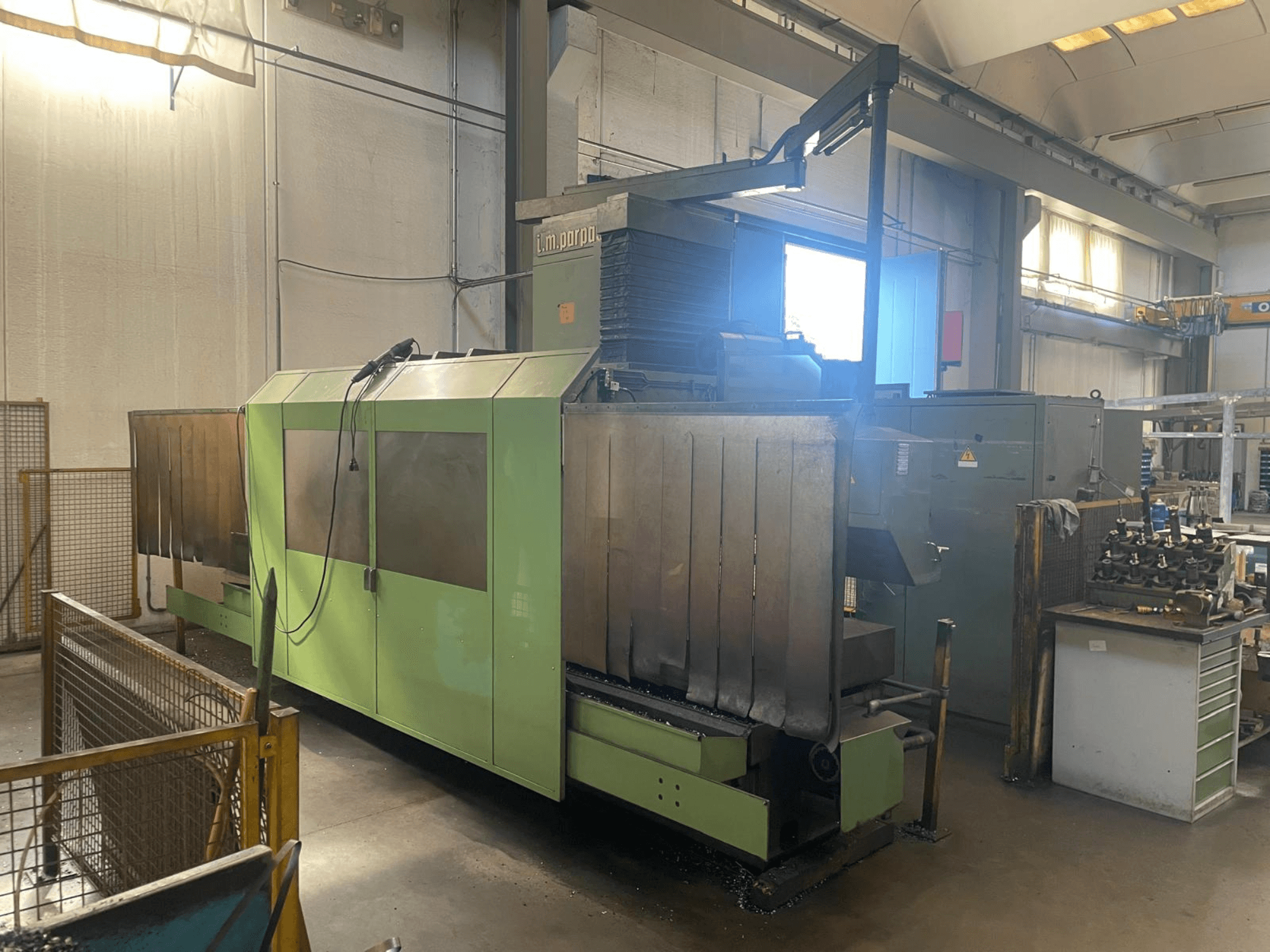 Görünür metal bileşenlere sahip yeşil endüstriyel makine, önden görünüm; kontrol paneli, güvenlik uyarıları ve yakınında iş istasyonu bulunmaktadır.