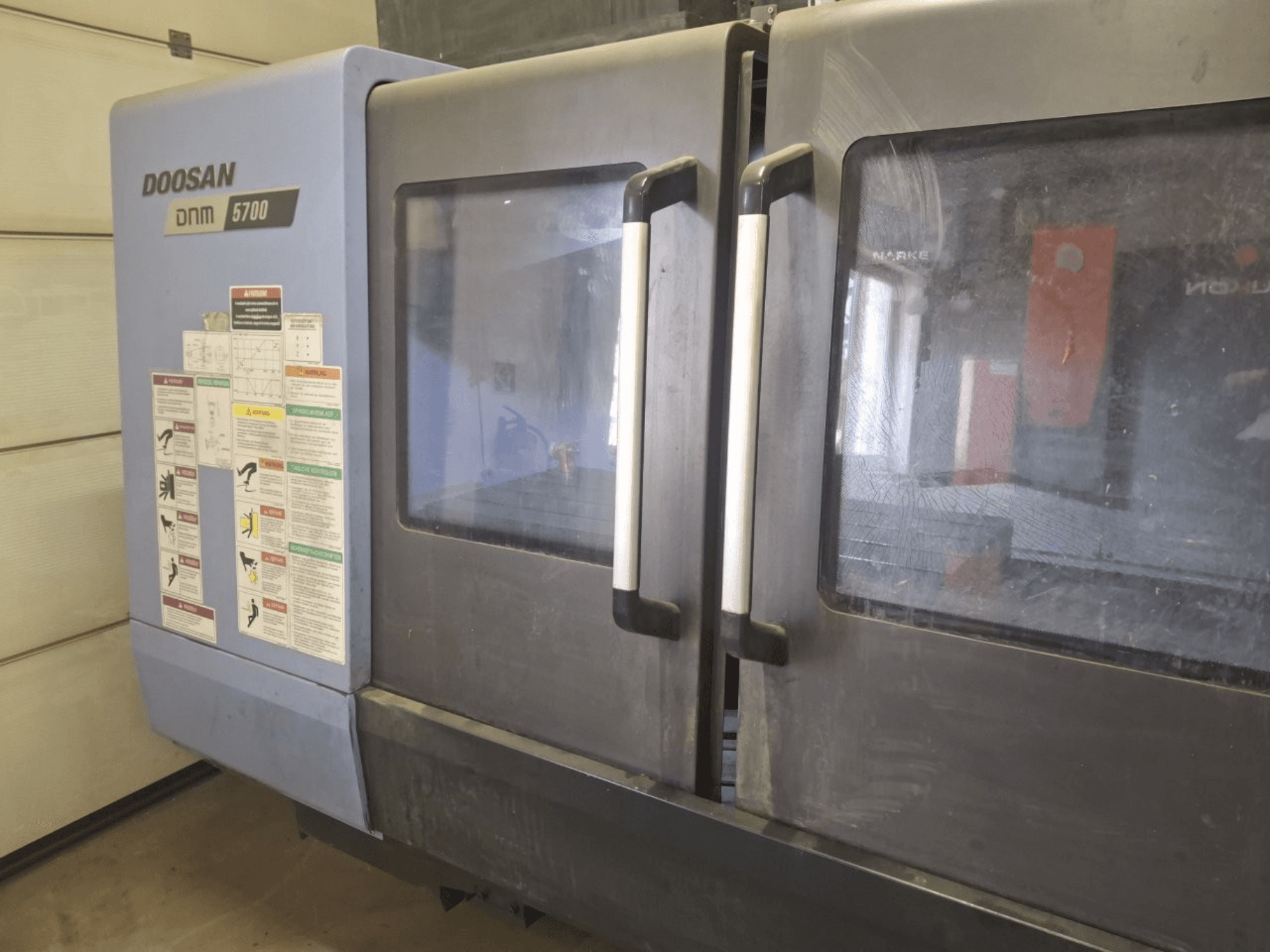 DOOSAN DNM 5700 Dik İşleme Merkezinin önden görünümü, çift kapı ve çalışma yönergelerini gösteren kontrol panelleri.