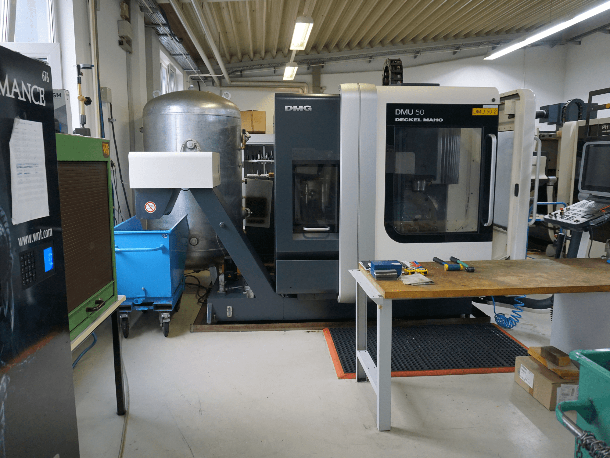 DMG DMU 50 freze makinesi bir atölyede, önden görünüm, ahşap bir masa üzerinde aletler ve yanında mavi tekerlekli bir çöp kutusu.