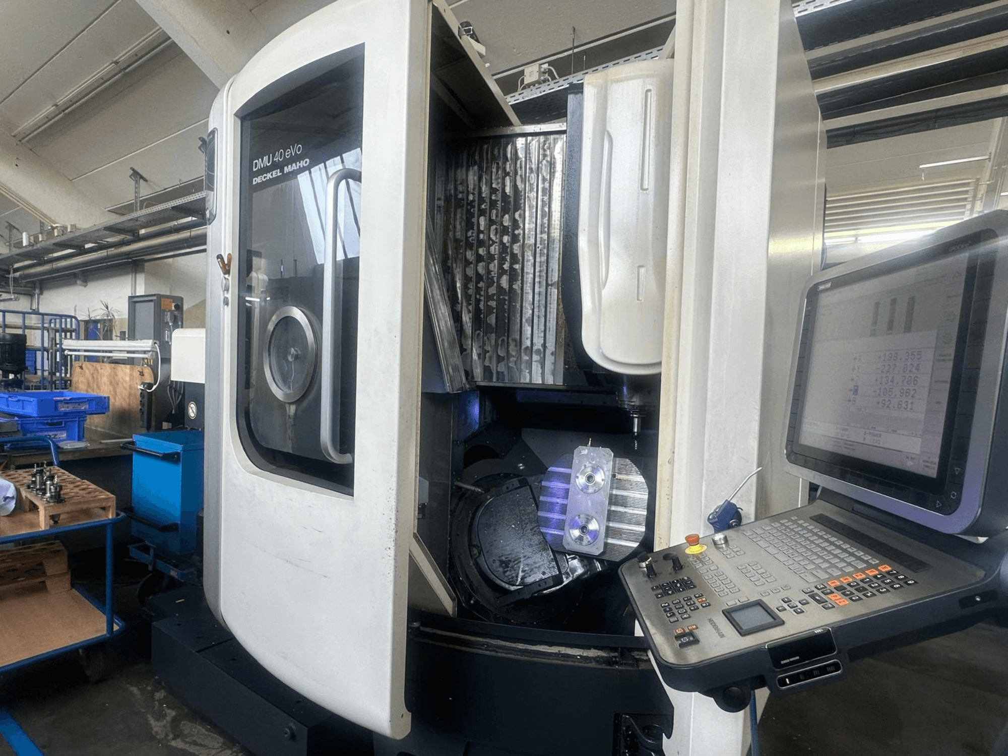 Deckel Maho'nun DMU 40 eVo freze makinesi bir atölyede, önden görünüm, kontrol paneli ve iş milindeki metal parçayı gösteriyor.