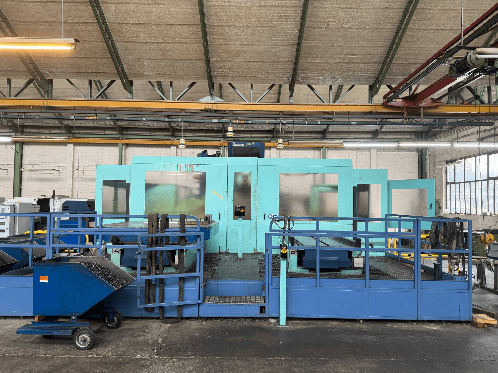 Koruyucu panelli endüstriyel mavi CNC makinesi, önden görünüm, alet deposu ve yan tarafta atık toplama arabası.