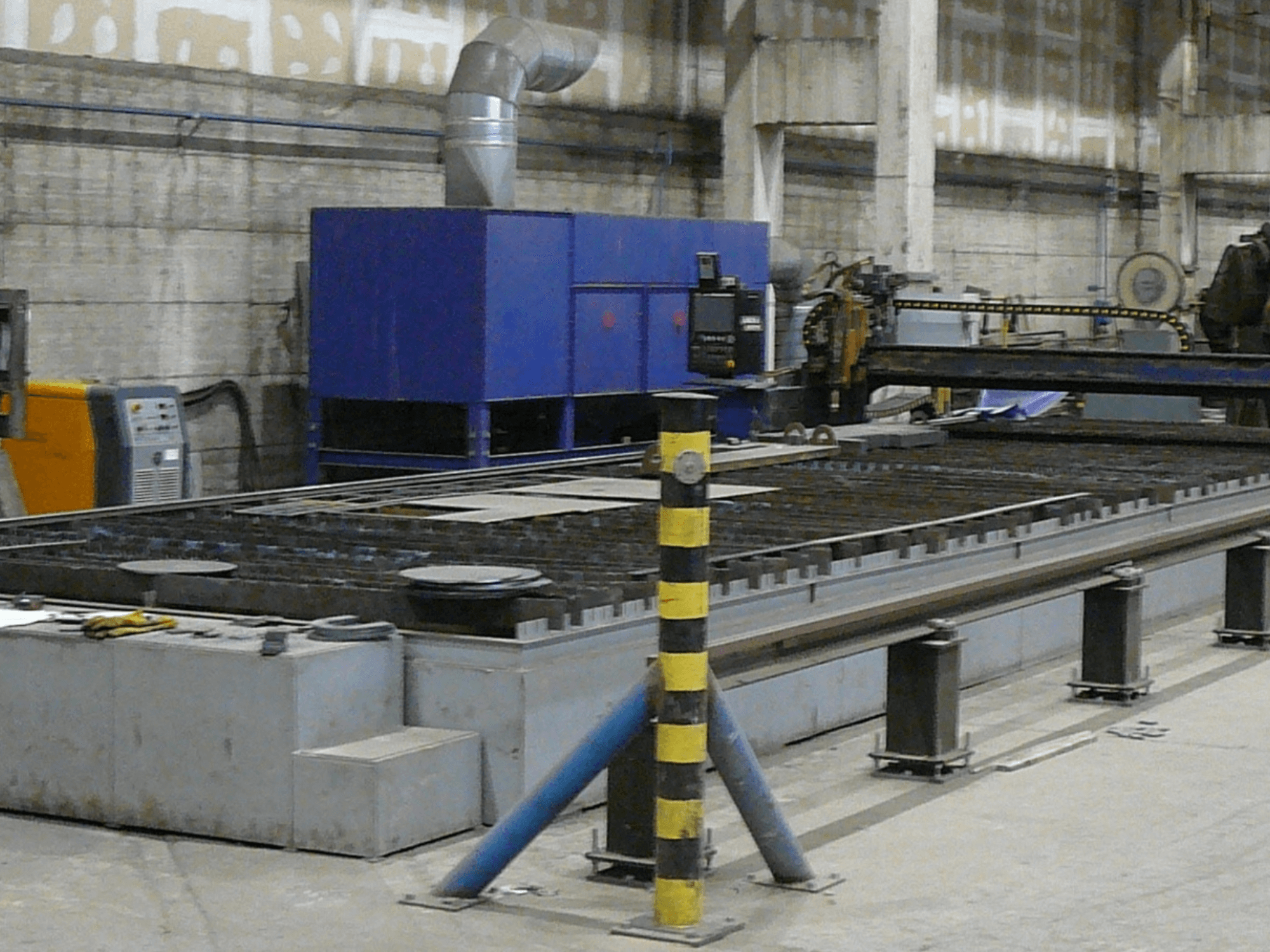 Endüstriyel metal kesme makinesi kurulumu, yandan görünüm, mavi kontrol kutusu, güvenlik şeritleri ve metal çalışma yüzeyi.