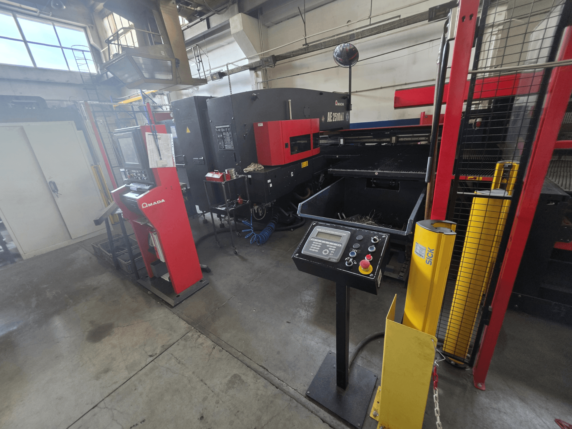 AMADA AE-2510NT CNC Delme Makinesi, kontrol paneli, güvenlik özellikleri ve çevresindeki endüstriyel ortamın görüldüğü önden görünüm.