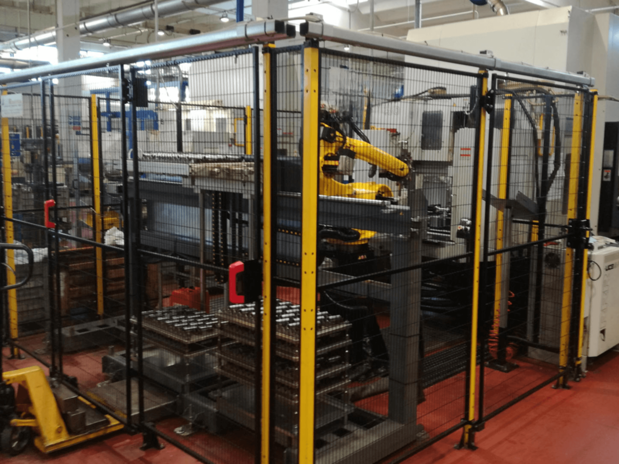 Enshu EV450 dikey işleme merkezi, robotik kollar ve çoklu istiflenmiş iş parçaları içeren çitle çevrili bir çalışma alanında. Önden görünüm.
