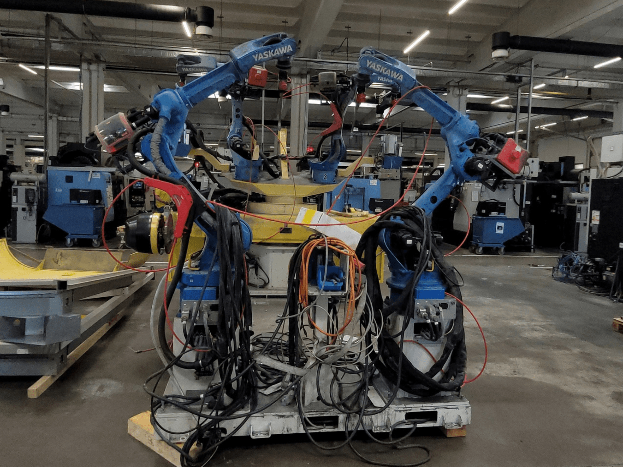 Yaskawa Robotik Kaynak Hücresi RWC L1_OS 15330, önden görünüm; uzun kablolara ve kaynak aletlerine sahip iki mavi robotun yer aldığı.