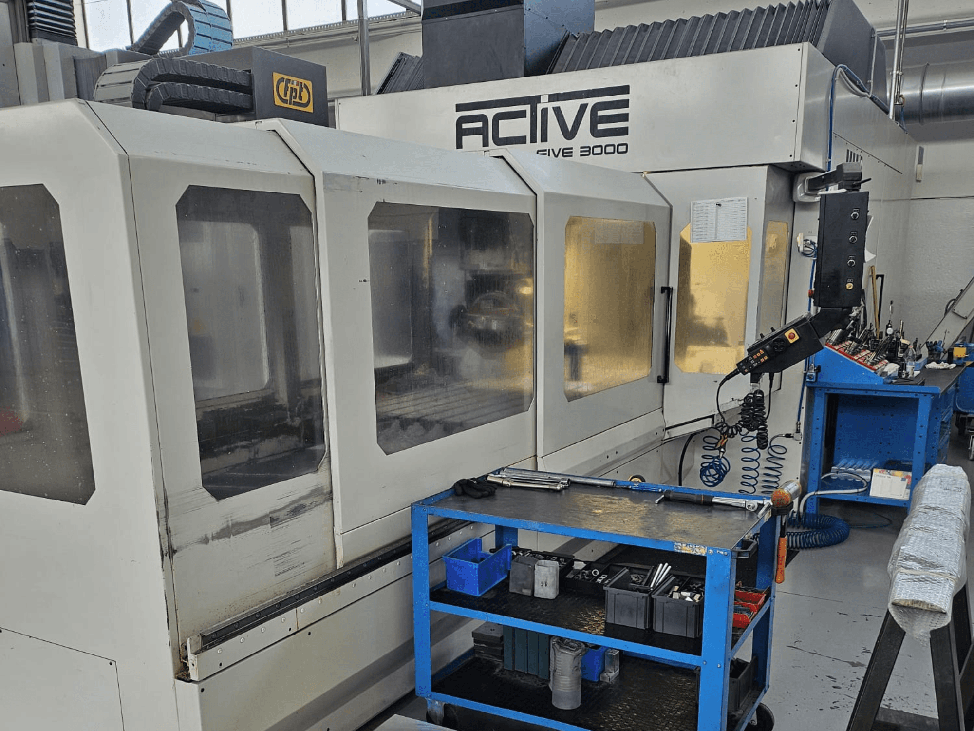 Bir atölyede aktif 3000 CNC makinesi, mavi bir araba üzerindeki aletlerle önden görünüm, görünür kontroller ve içerideki çalışma alanı.