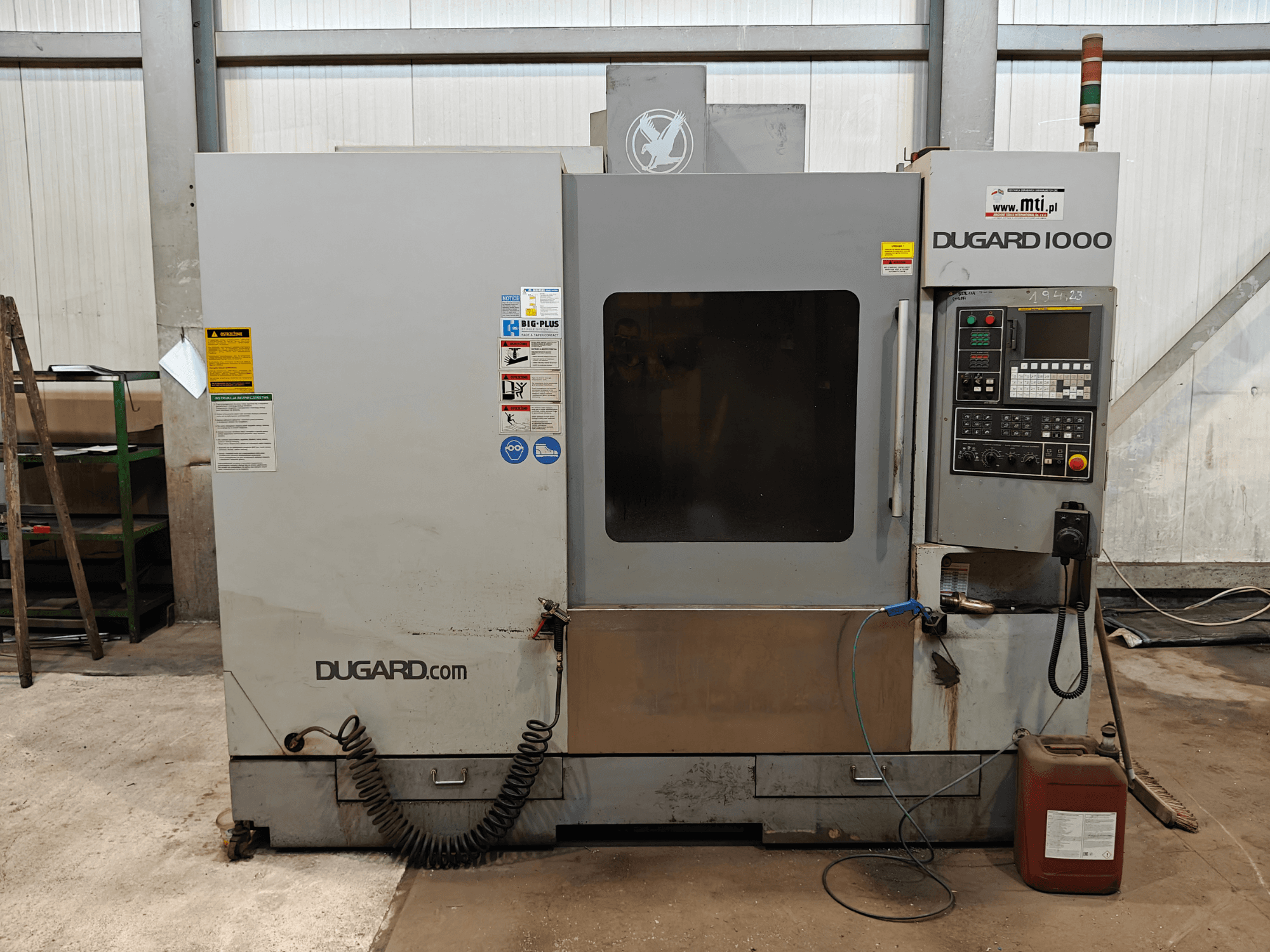 Dugard 1000 CNC makinesi, önden görünüm, kontrol panelini, güvenlik etiketlerini ve yağ kabını gösterir, atölye ortamında.