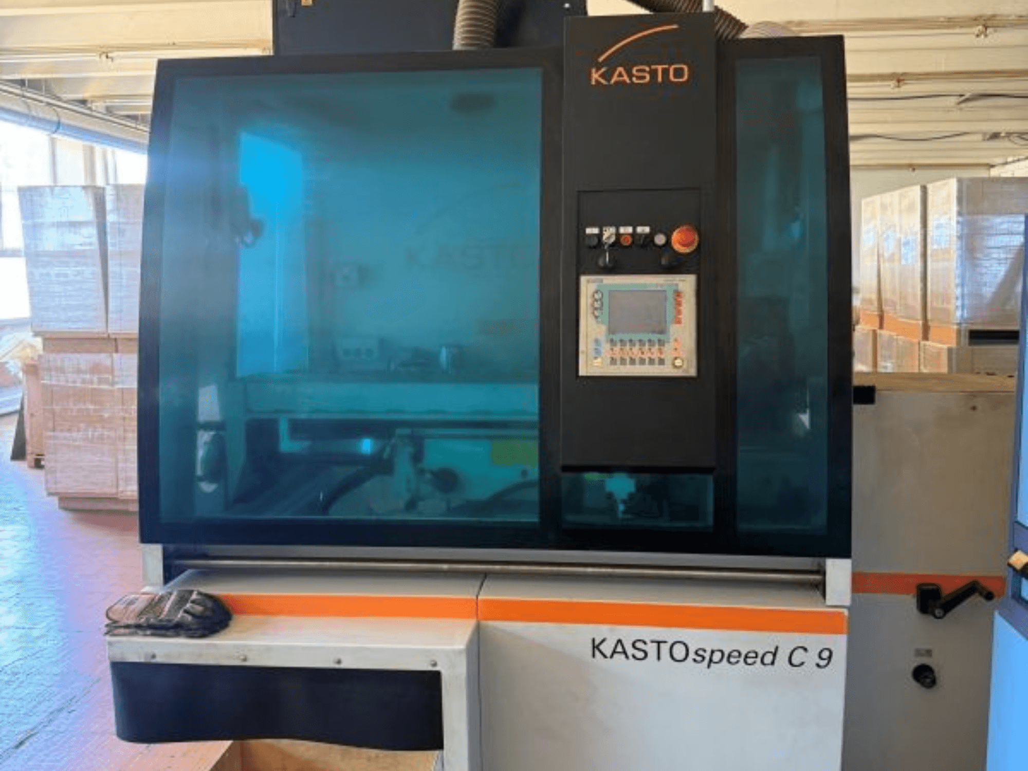 KASTO KASTOspeed C 9 dairesel metal testeresinin önden görünümü; kontrol paneli ve güvenlik camı göze çarparken, arka planda ahşap kasalar yer almaktadır.