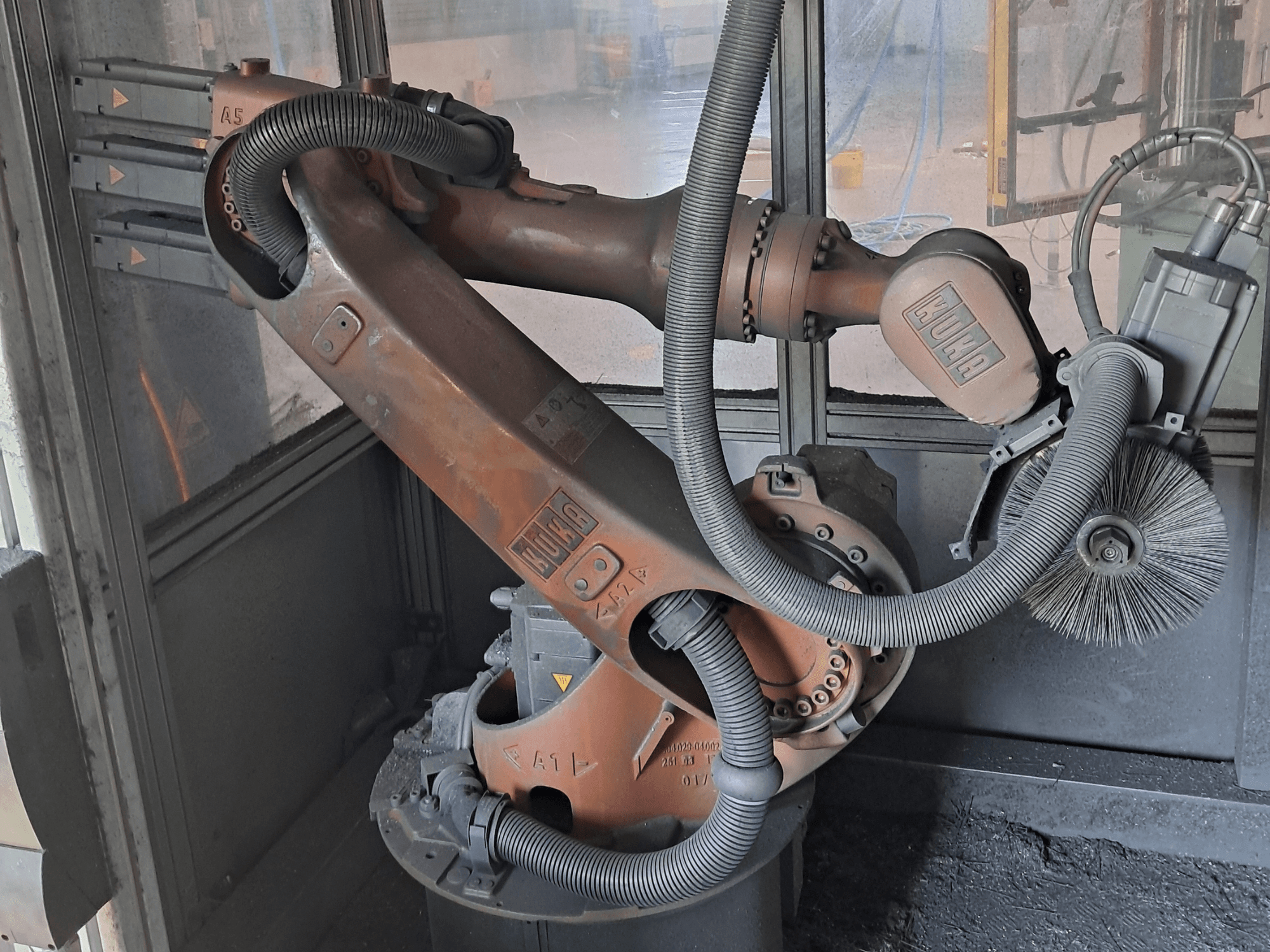 Bir çalışma ortamında yer alan KUKA KR 16-2 robot kolunun yandan görünümü; kablolar, fırçalar ve mekanik eklemler açıkça görülmektedir.
