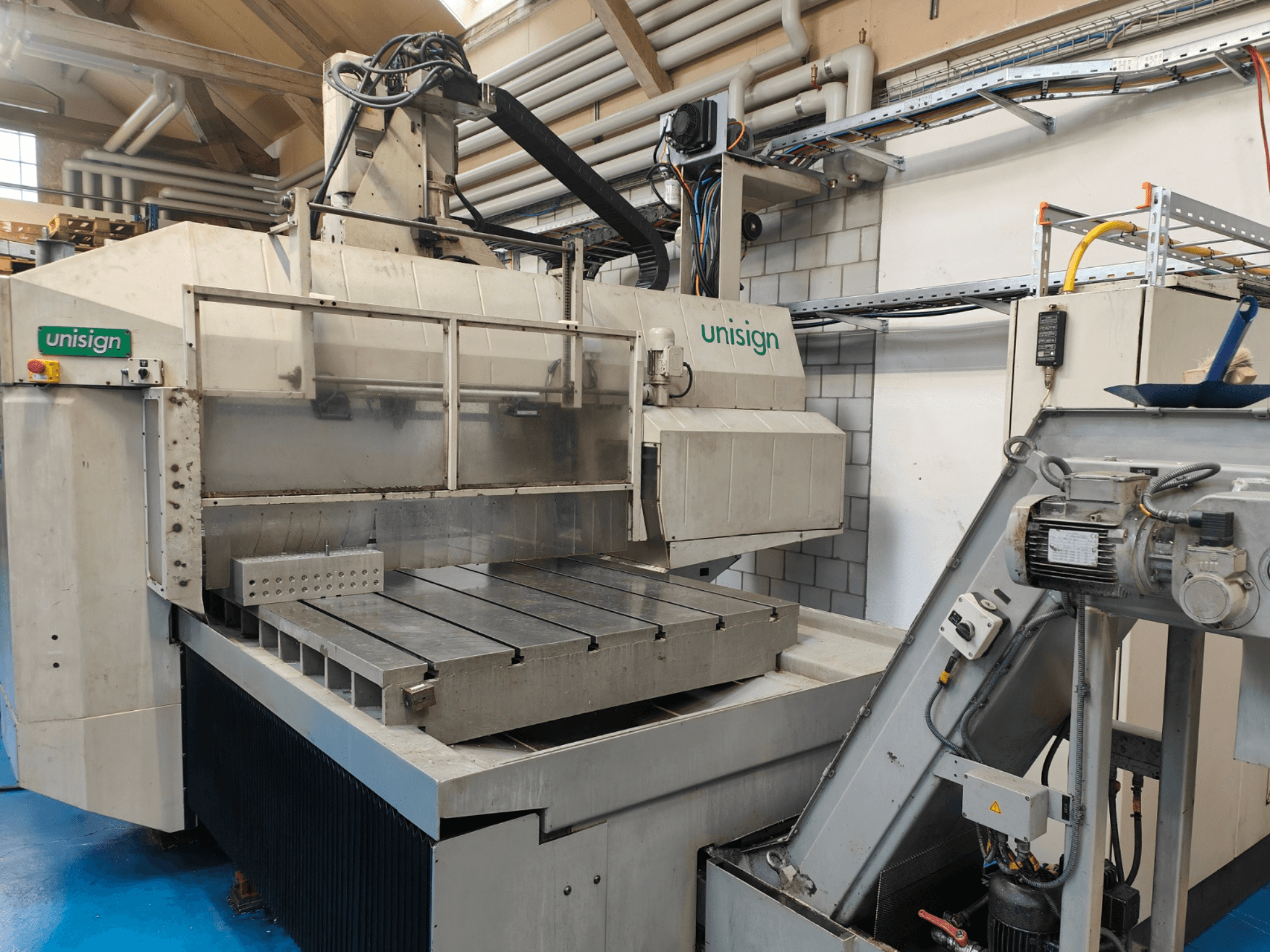 Bir atölyede Unisign CNC makinesi, önden görünüm, büyük bir masa, kontrol paneli ve endüstriyel tasarım öğeleri.