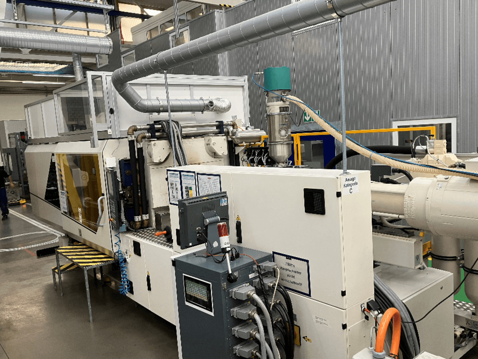 Enjeksiyon kalıplama makinesi, önden görünüm, fabrika ortamında marka etiketi, kontrol paneli ve görünür hortumlar.