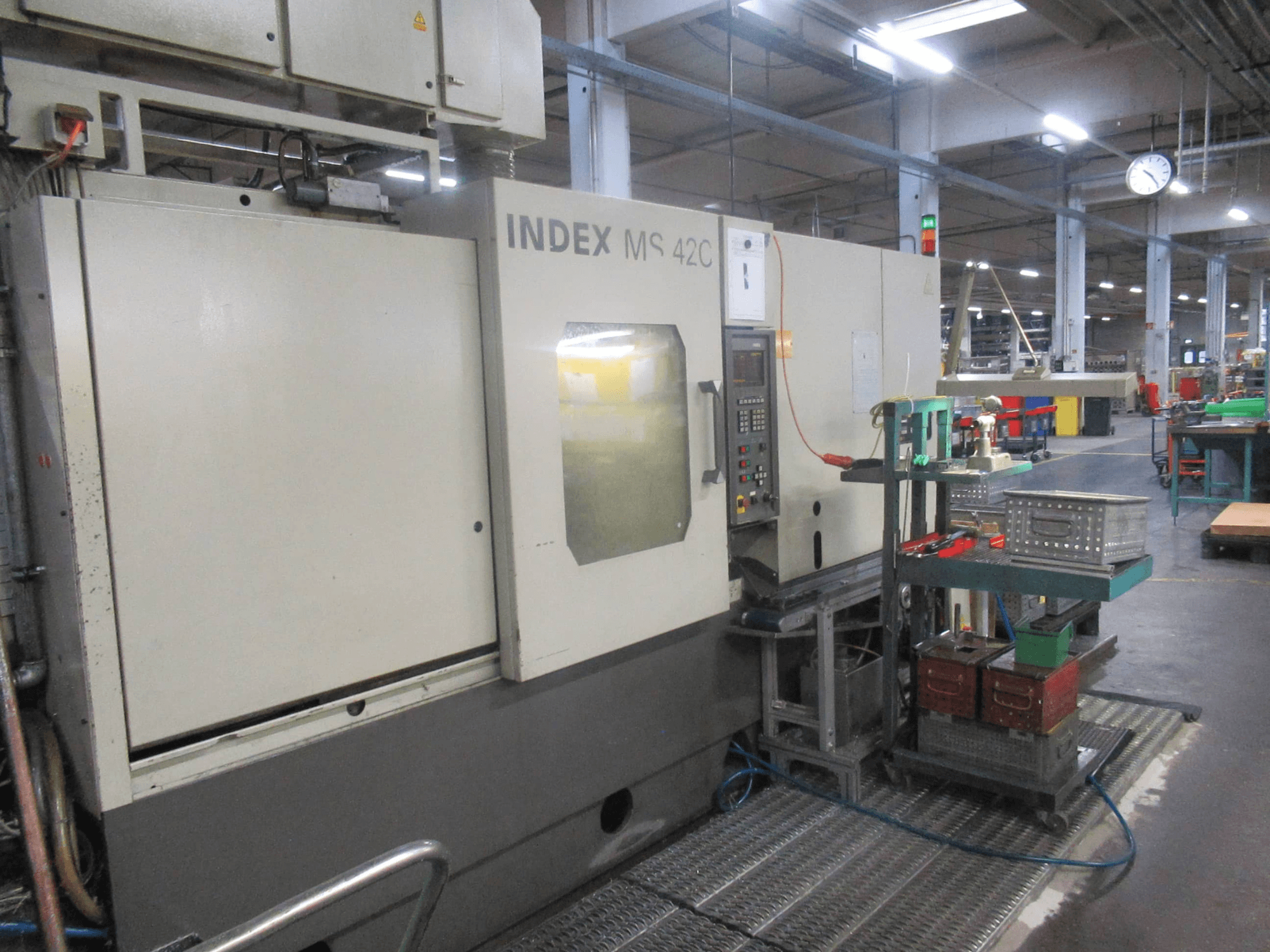 INDEX MS 42C CNC makinesi bir atölyede, kontrol panelini ve tekerlekli araba üzerindeki alet deposunu gösteren yandan görünüm.