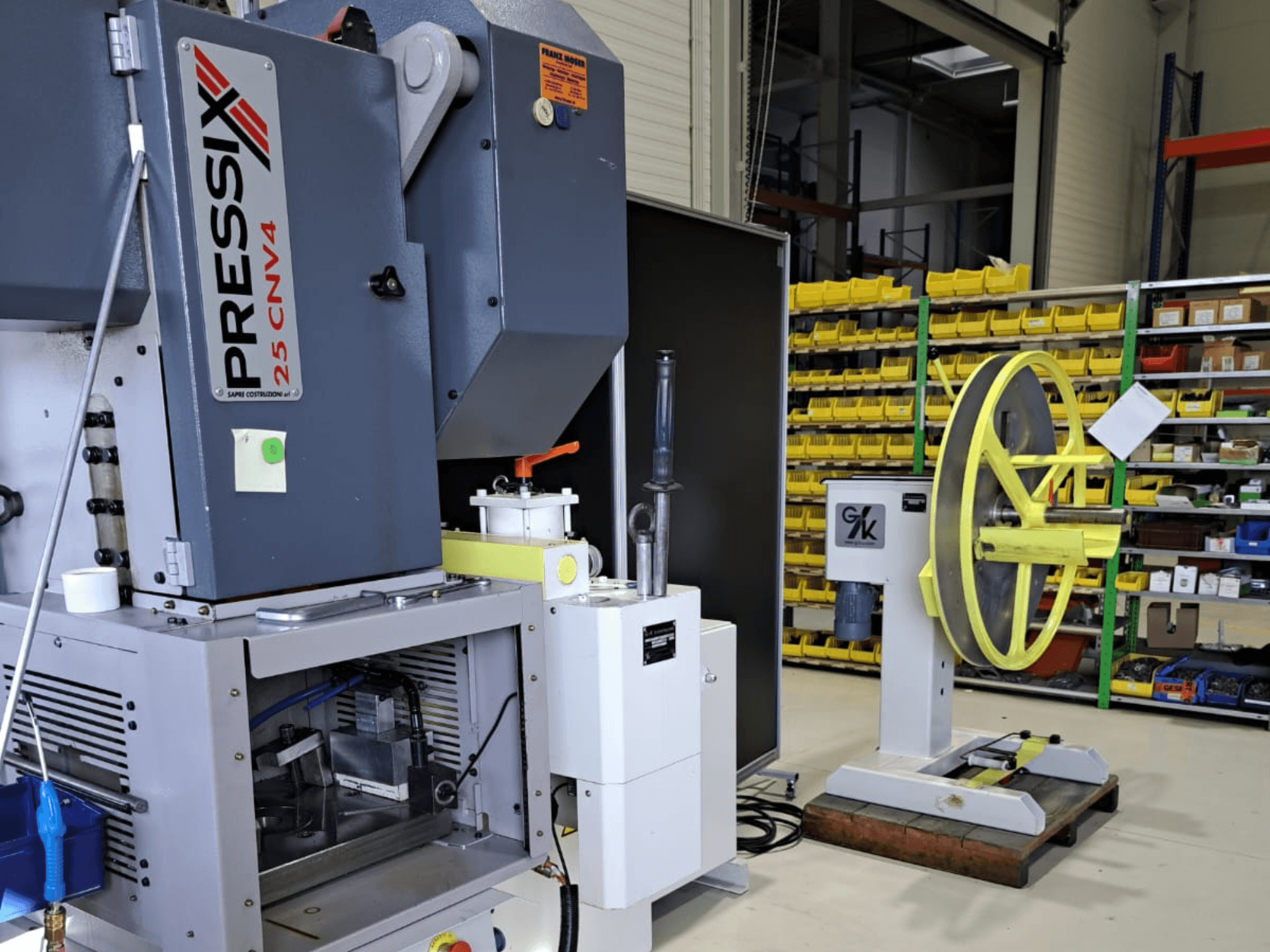 Solda PRESSIX 25 CNV4 ve sağda gri bir taban üzerine monte edilmiş sarı bir tekerlek içeren endüstriyel makine kurulumu, önden görünüm.
