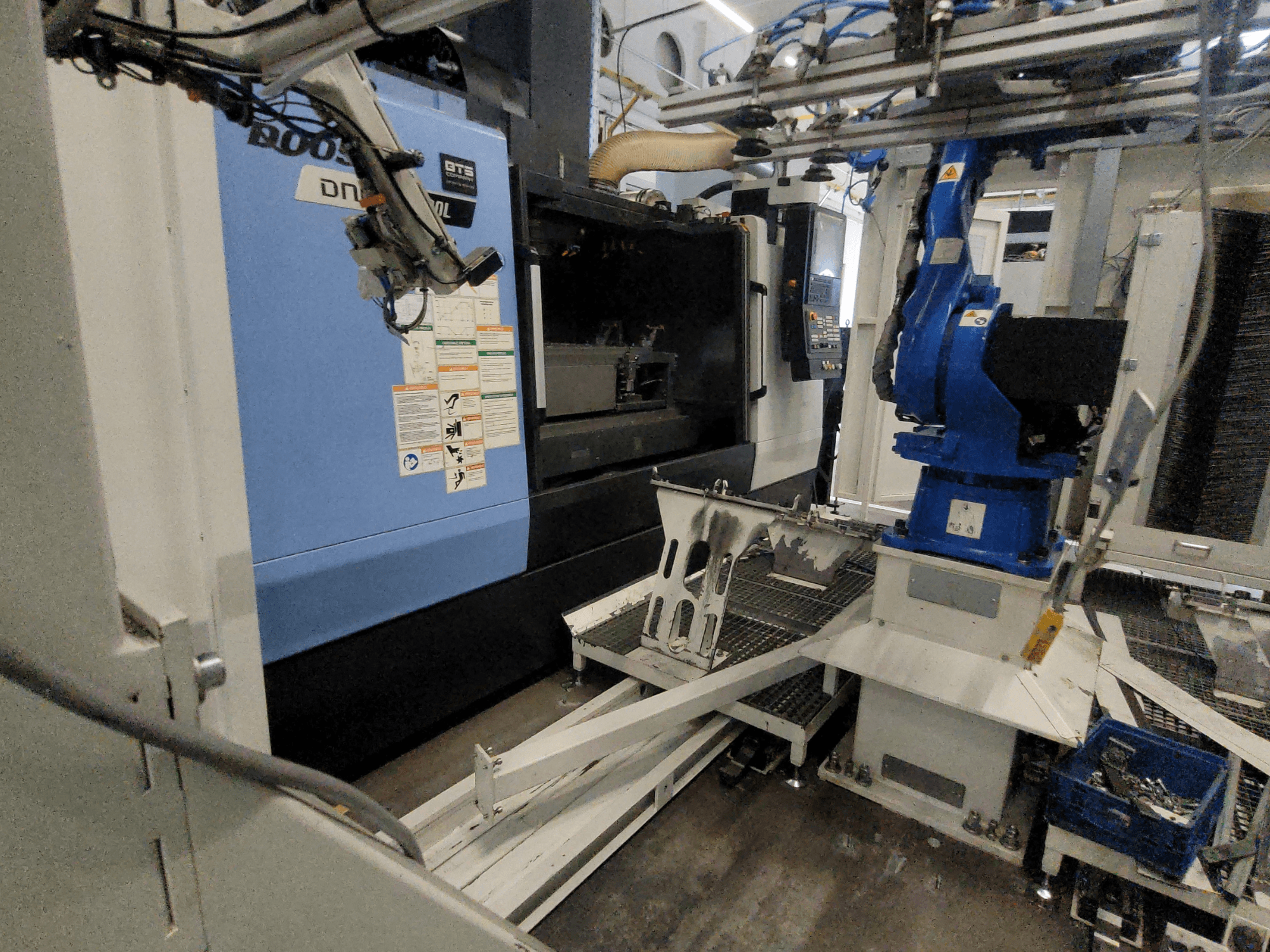 DOOSAN DNM 6700L Dikey İşleme Merkezi’nin yan görünümü; robot kolu ve kontrol paneli görülebilir ve makinenin kurulumunu göstermektedir.