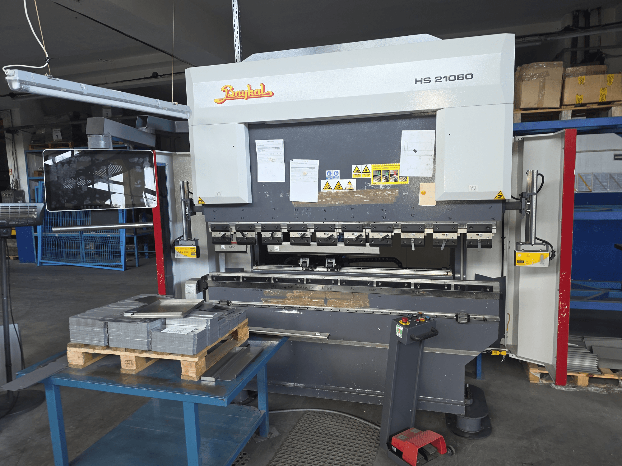 Bir atölyede bulunan Baykal APHS 21060 pres fren makinesi; kontrol paneli, üst üste istiflenmiş metal levhalar ve güvenlik işaretlerini gösteren yandan görünüm.