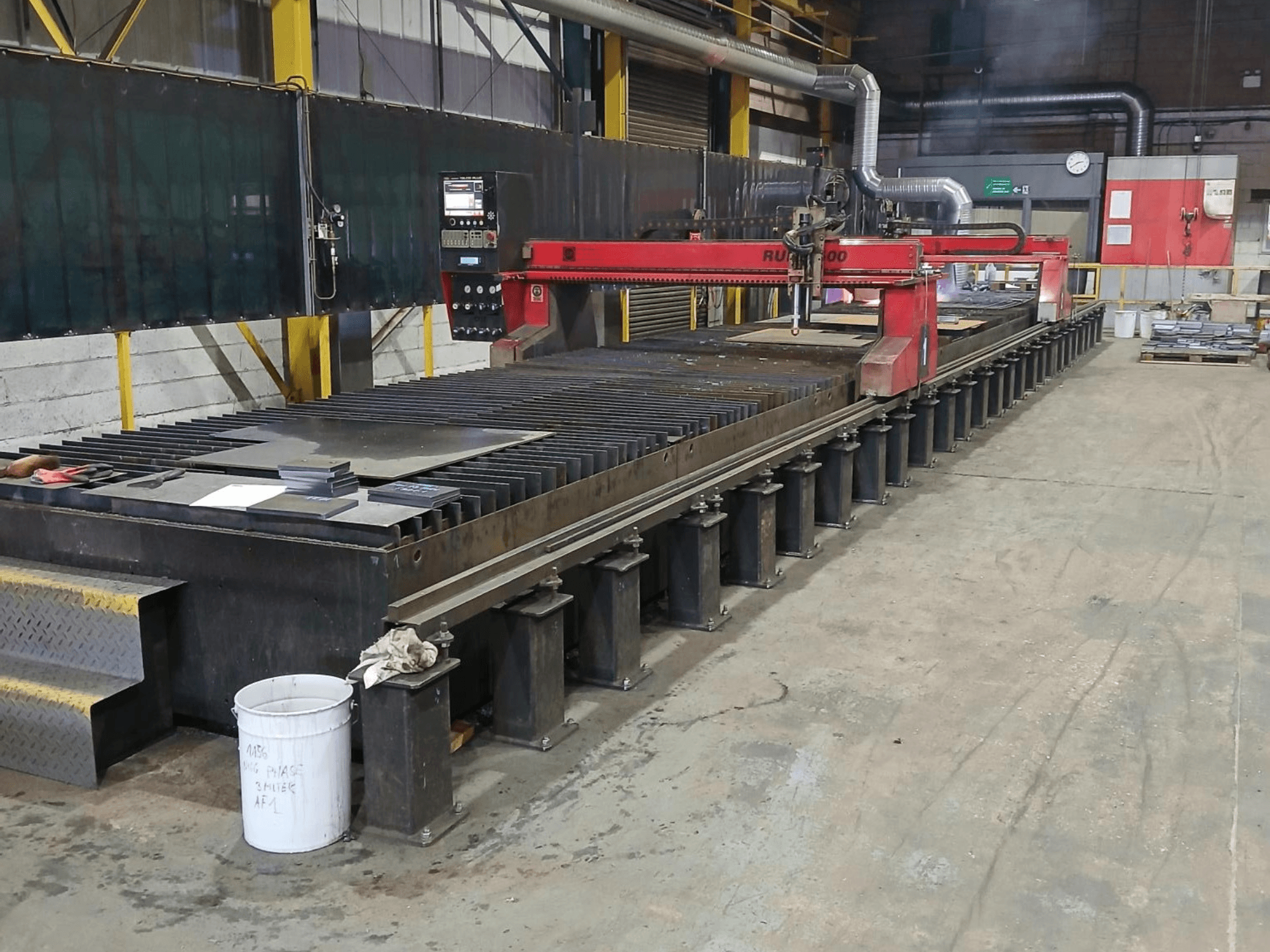 Kırmızı RUIDA 300 kesme makinesi, yandan görünüm, çelik bir masa üzerinde metal levha ile, endüstriyel bir çalışma alanında yer almaktadır.