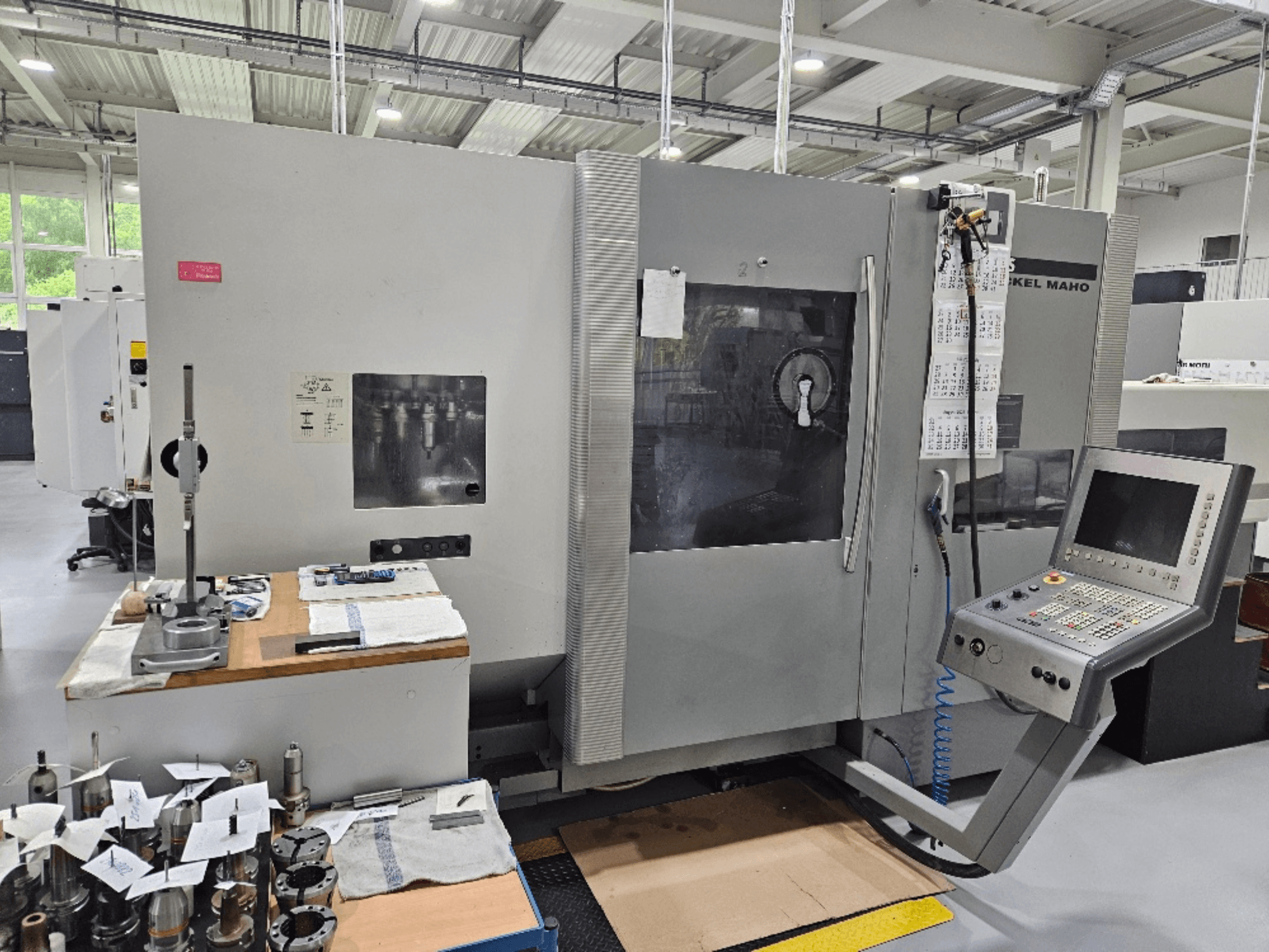 Bir DMG Mori DMU 70 makinesinin önden görünümü, kontrol paneli, ahşap bir masa üzerinde aletler ve yanında çeşitli iş parçaları.