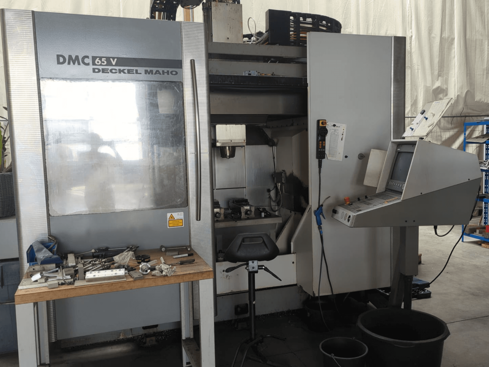 Kontrol paneli, tezgah üzerindeki takımlar ve çalışma alanı düzeniyle birlikte bir DECKEL MAHO DMC 65 V dikey işleme merkezinin önden görünümü.