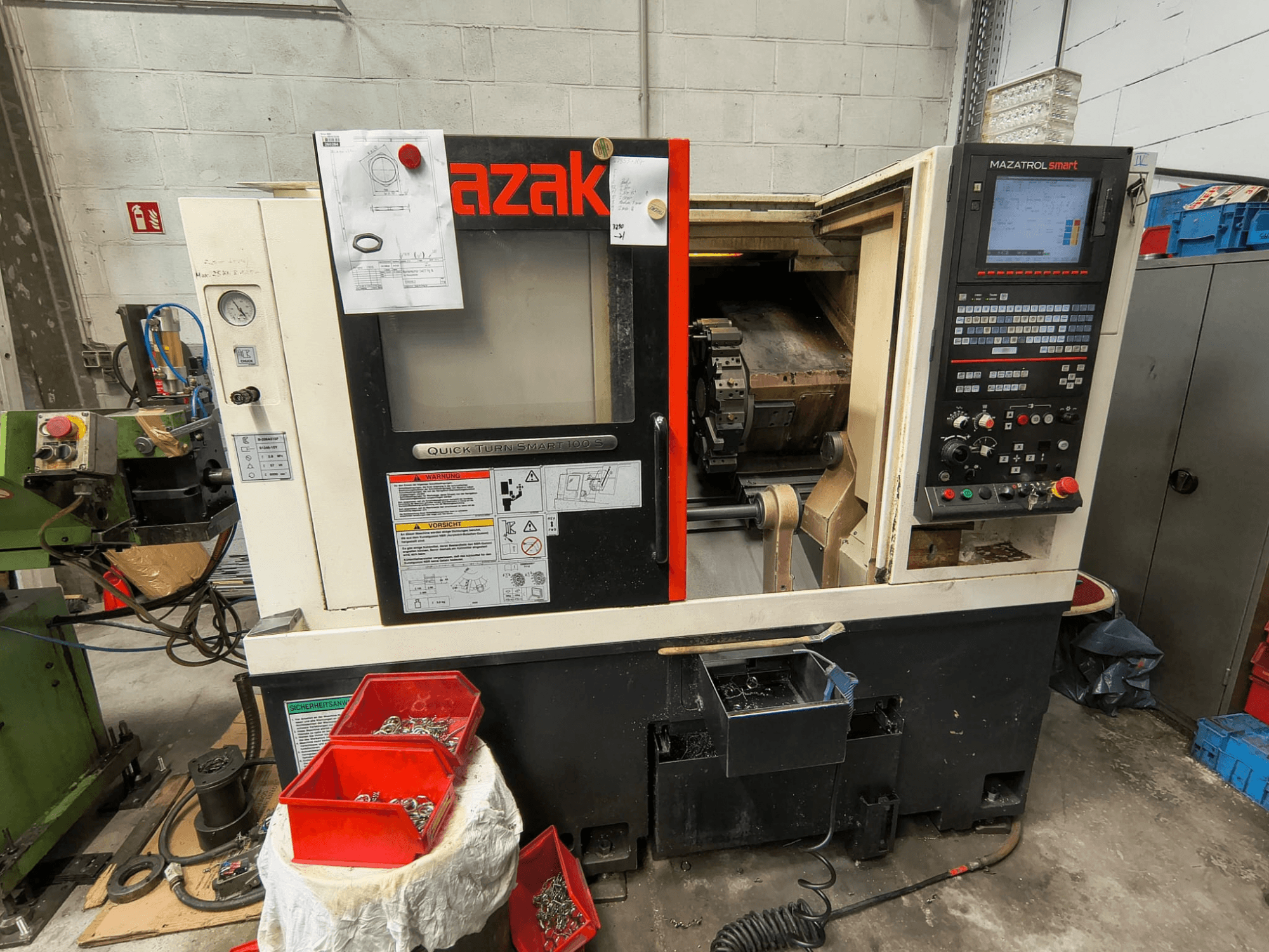 Mazak Quick Turn Smart 100 S, kontrol paneli, soğutma sıvısı tepsisi ve vidaların bulunduğu saklama bölmelerini gösteren yandan görünüm.