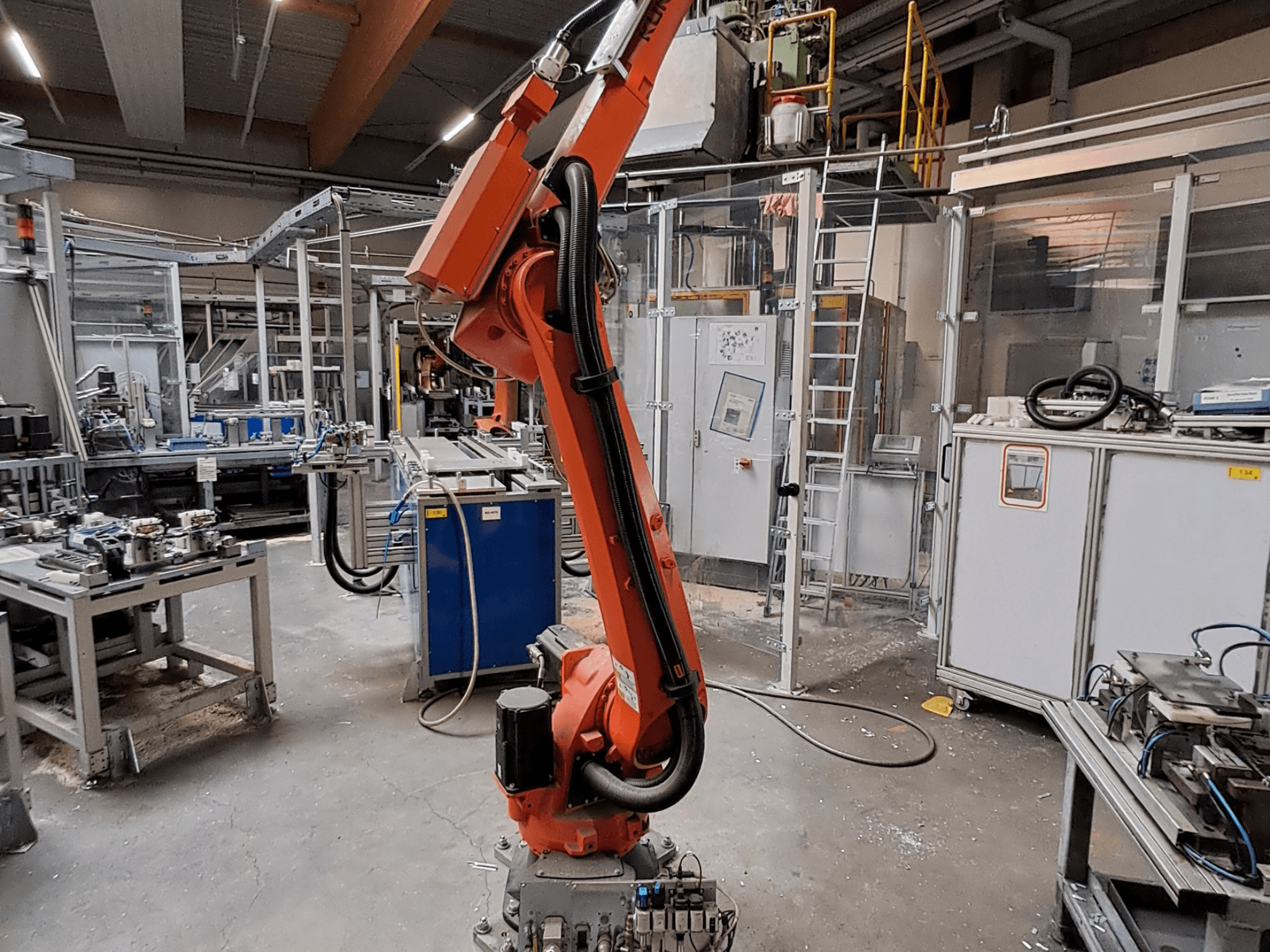 Endüstriyel bir ortamda bulunan KUKA KR 26 R3100/SEL robot kolu; arka planda çalışan ekipmanların da görüldüğü bir yan görünüm.