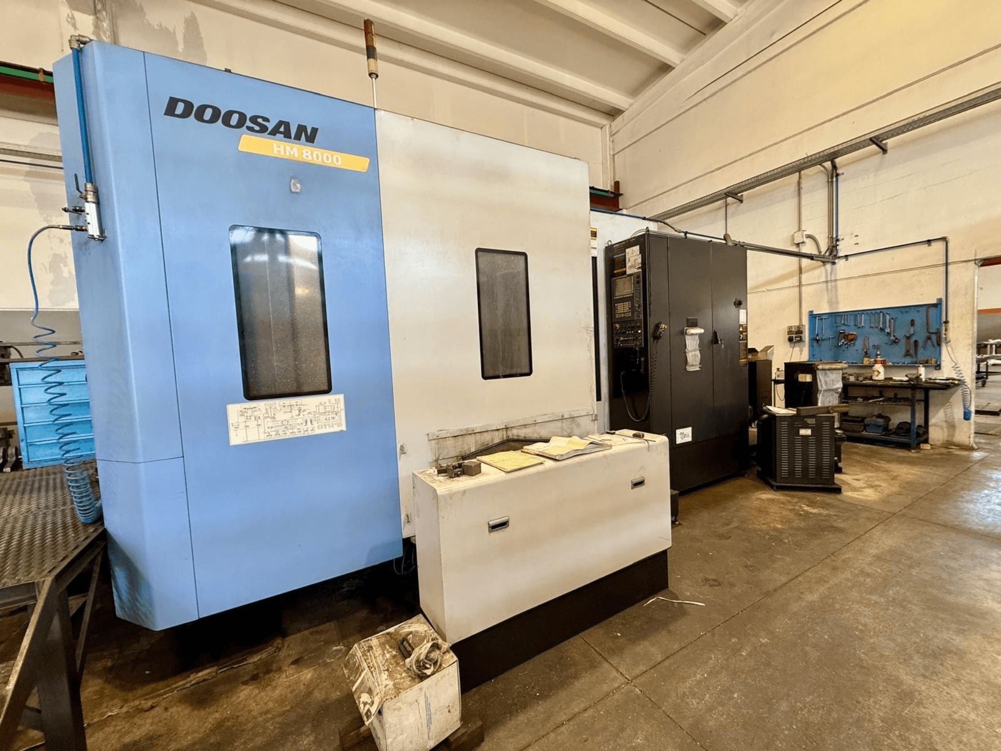 Mavi renkli DOOSAN HM 8000 yatay işleme merkezi, önden görünüm; kontrol paneli ve çevresindeki atölye ekipmanları görülmektedir.