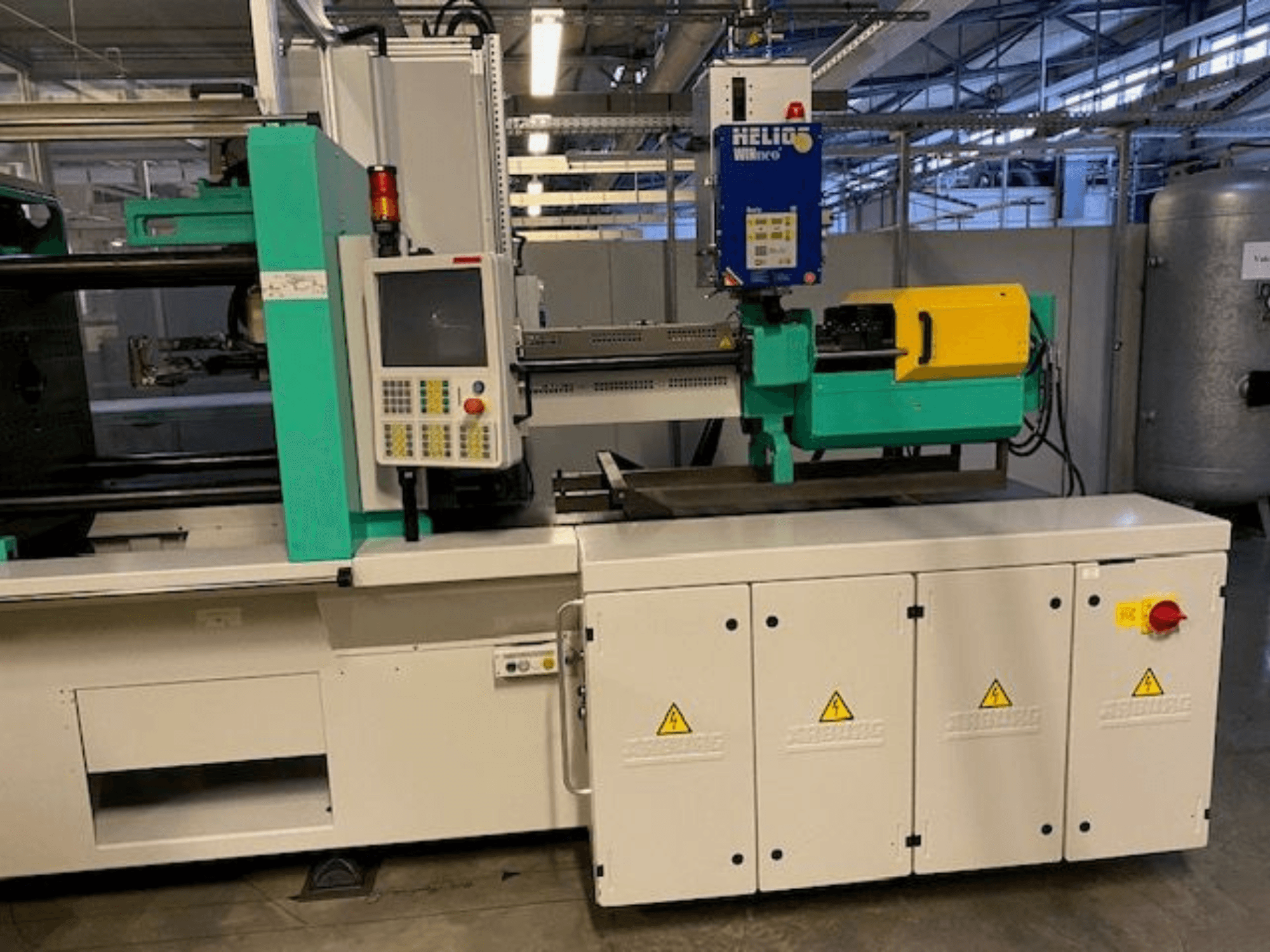 Endüstriyel enjeksiyon kalıplama makinesi, model HELIO, önden görünüm, kontrol paneli, güvenlik korumaları ve acil durdurma düğmesi.
