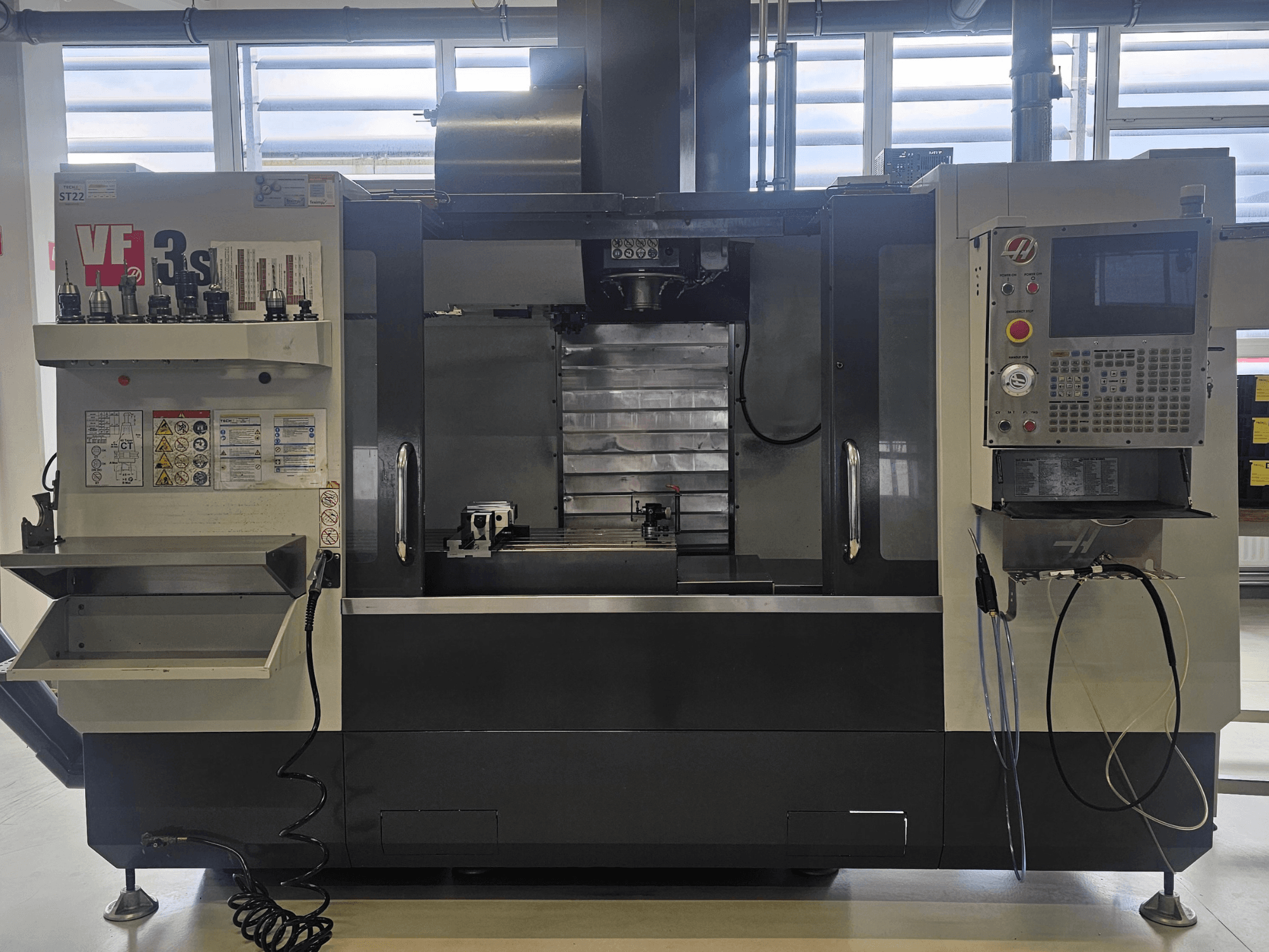 Haas VF3SS CNC freze, önden görünüm, takım tutucular, kontrol paneli ve boş soğutma sıvısı tepsisi, güvenlik etiketleri görünür.
