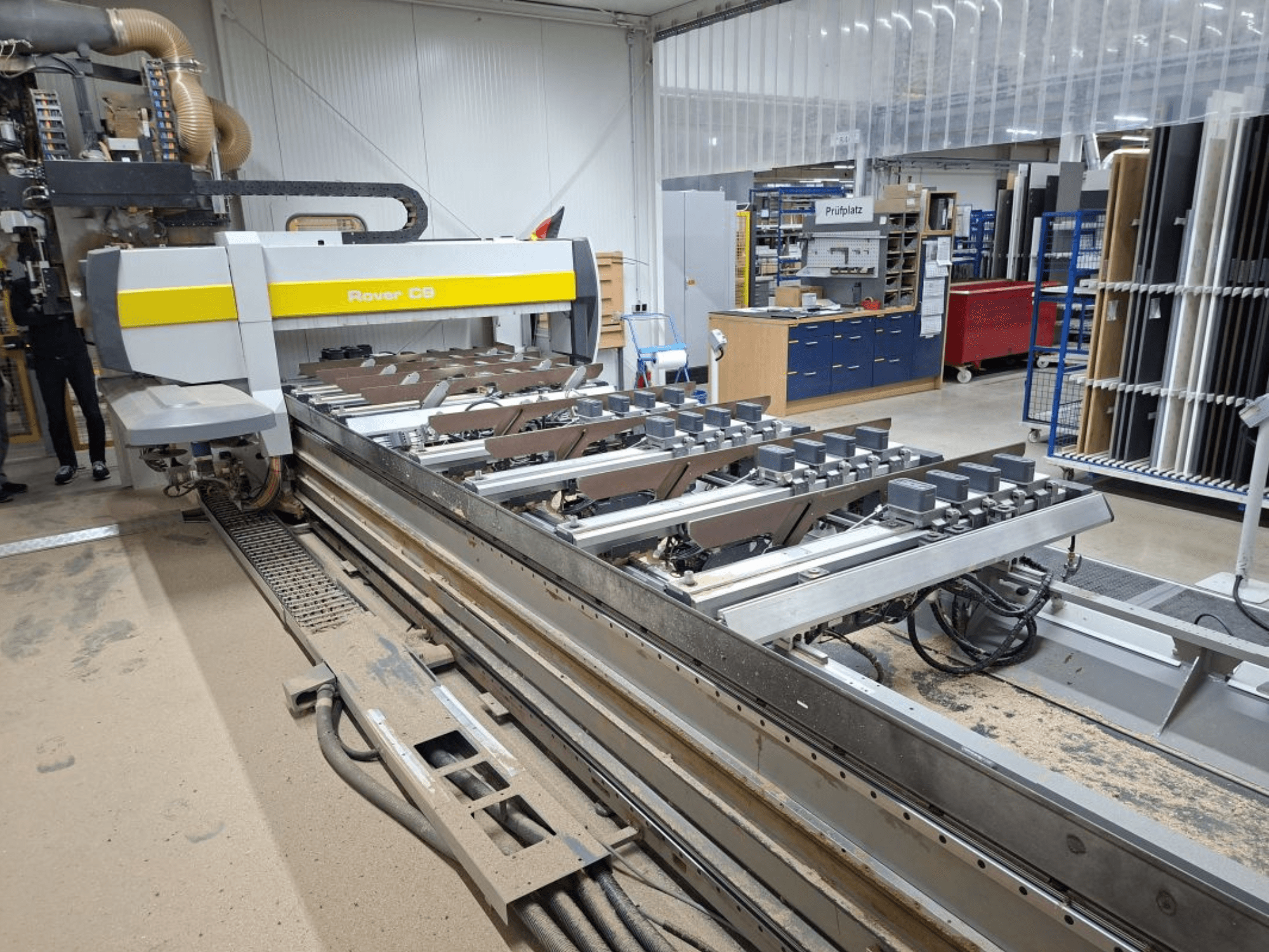 Bir atölyedeki Rover C9 CNC makinesi; önden görünüm, zeminde toz birikimi ile birlikte kesme takımlarını ve konveyör sistemini göstermektedir.