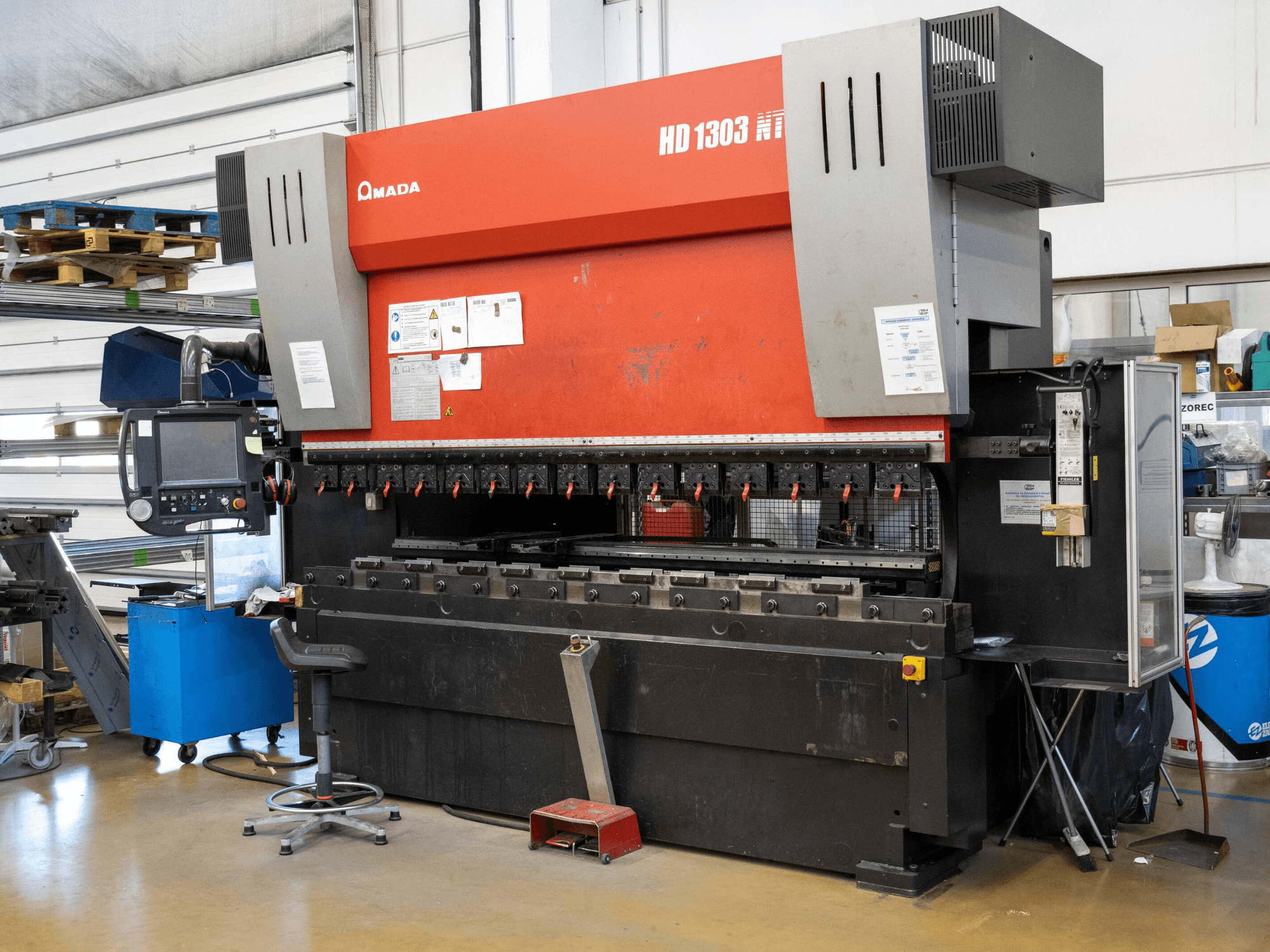 Amada HD 1303 hidrolik abkant pres atölyede, önden görünüm; çoklu kelepçeler, kontrol paneli ve ayarlanabilir tabure özellikleri.