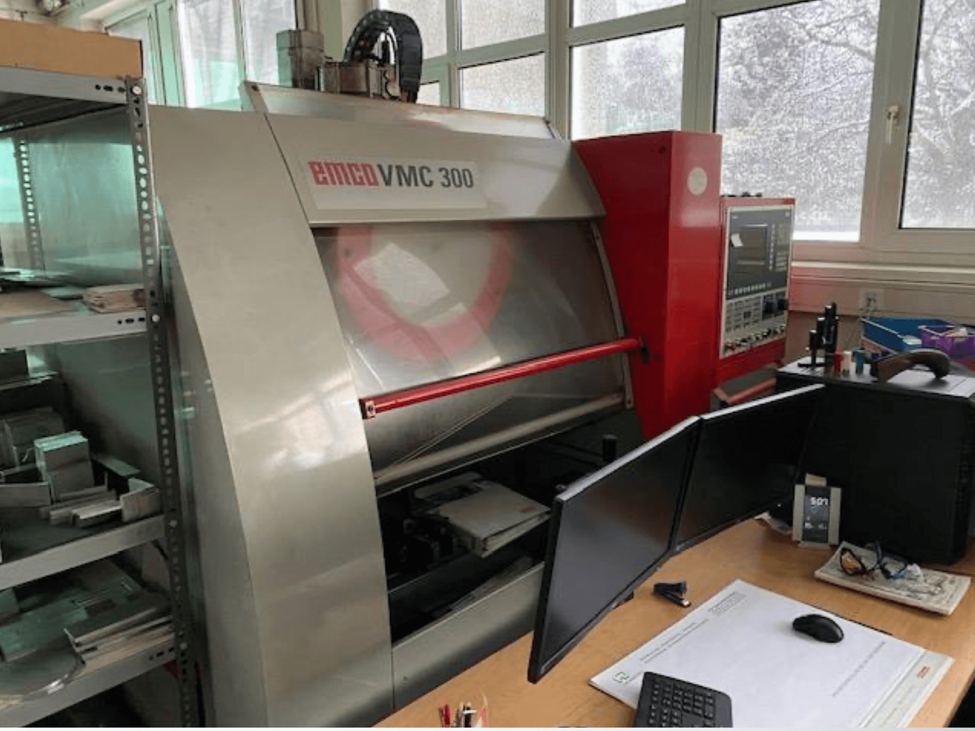 Bir atölyede bulunan EMCO VMC 300 dikey işleme merkezi; kontrol paneli ve çift monitörlü yakındaki çalışma istasyonunu gösteren yandan görünüm.