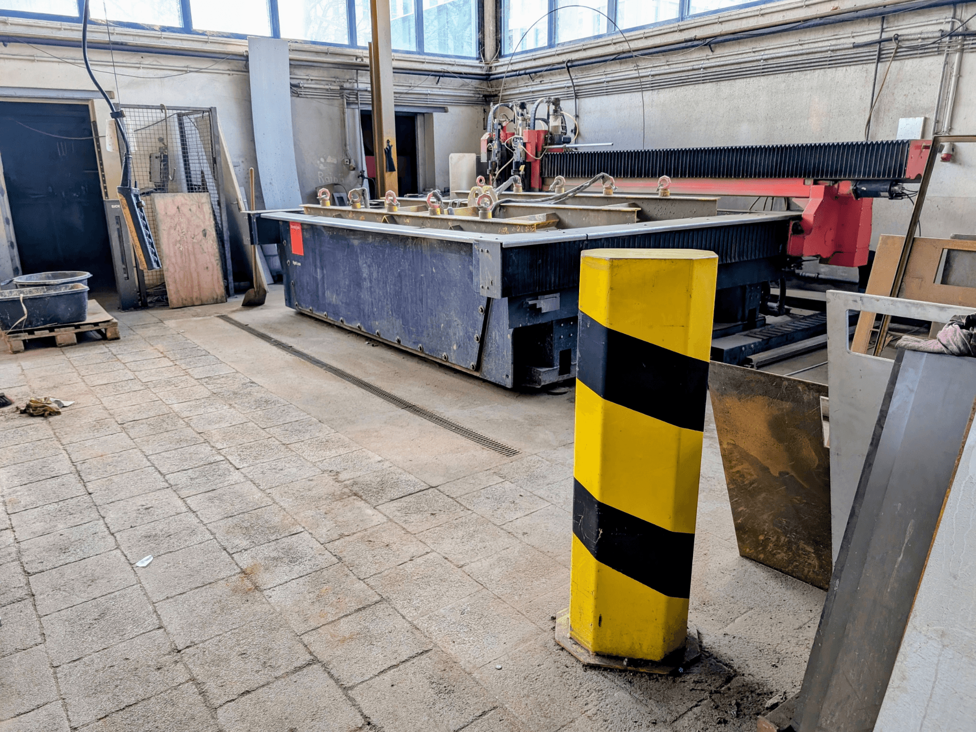 Bir atölyede Bystronic Byjet L 2030 su jeti kesim makinesi, yandan görünüm, sarı güvenlik direği ve çevresindeki malzemeler.