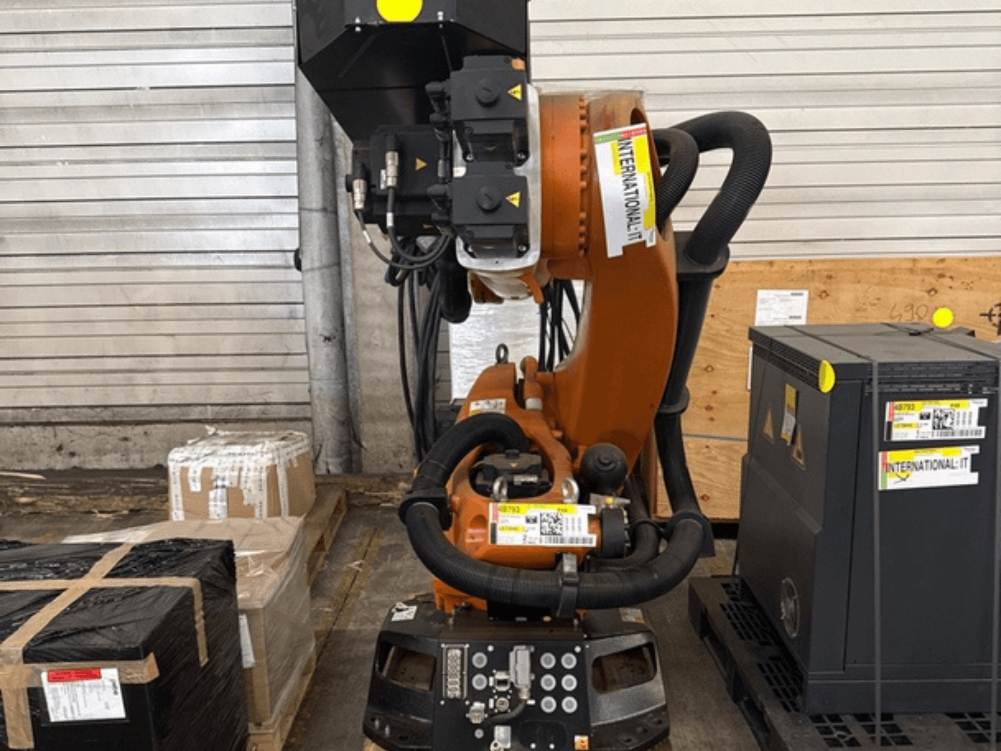 KUKA KR 210 R2700 yedek robot kolu, önden görünümde; paketlerin ve ekipmanların yanında bir palet üzerinde sergileniyor.