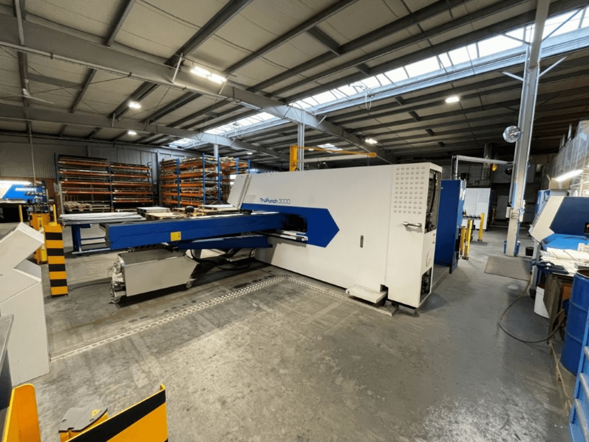 Fabrika ortamında çekilmiş CNC delme presi TRUMPF TruPunch 3000’in önden görünümü, mavi ve beyaz renkli tasarımını ve çalışma bileşenlerini gözler önüne seriyor.