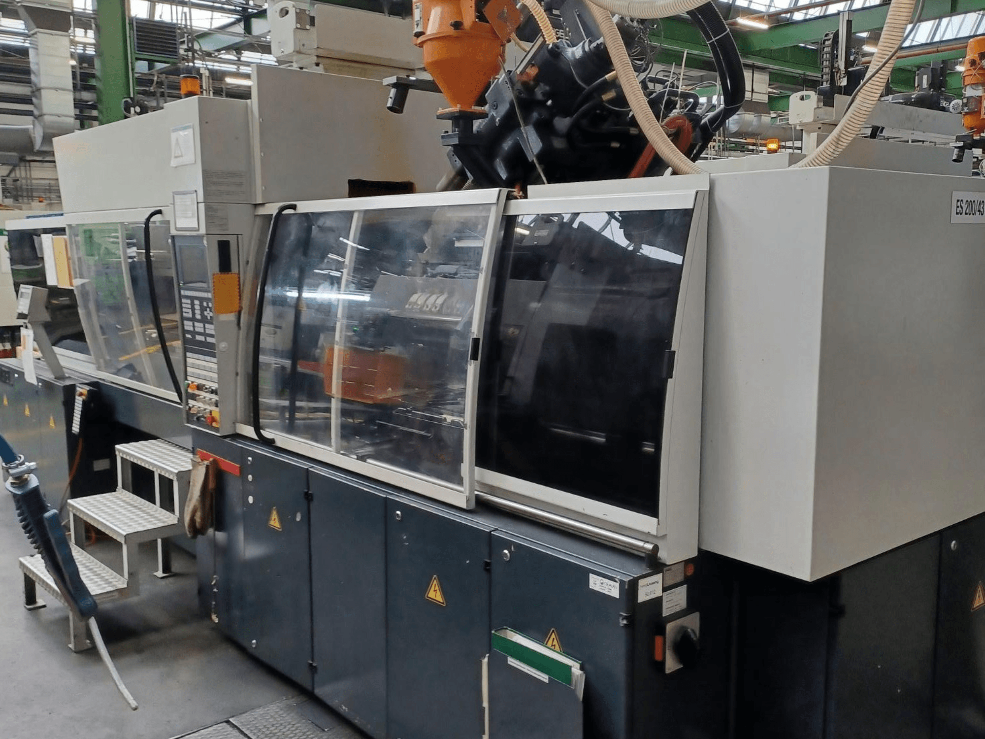 Markası ve modeli bilinmeyen, şeffaf güvenlik kapakları ve kontrol paneli bulunan bir plastik enjeksiyon makinesinin önden görünümü.