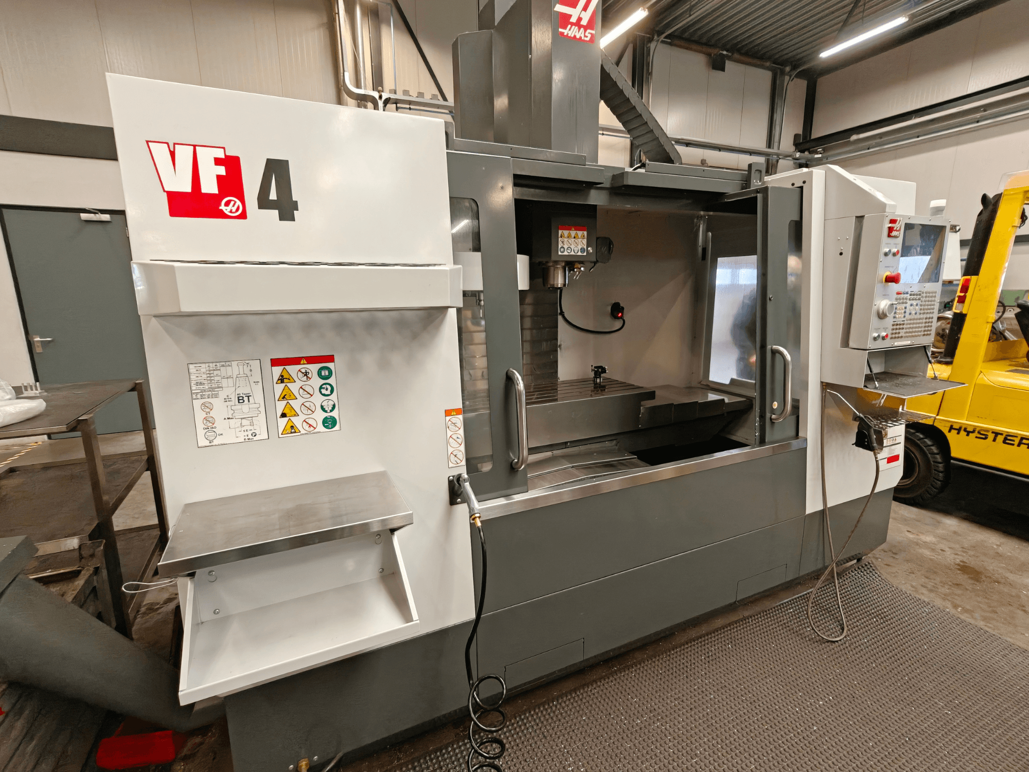 HAAS VF-4 dikey işleme merkezi, önden görünüm, kontrol paneli, çalışma alanı ve yan taraftaki güvenlik etiketleri.