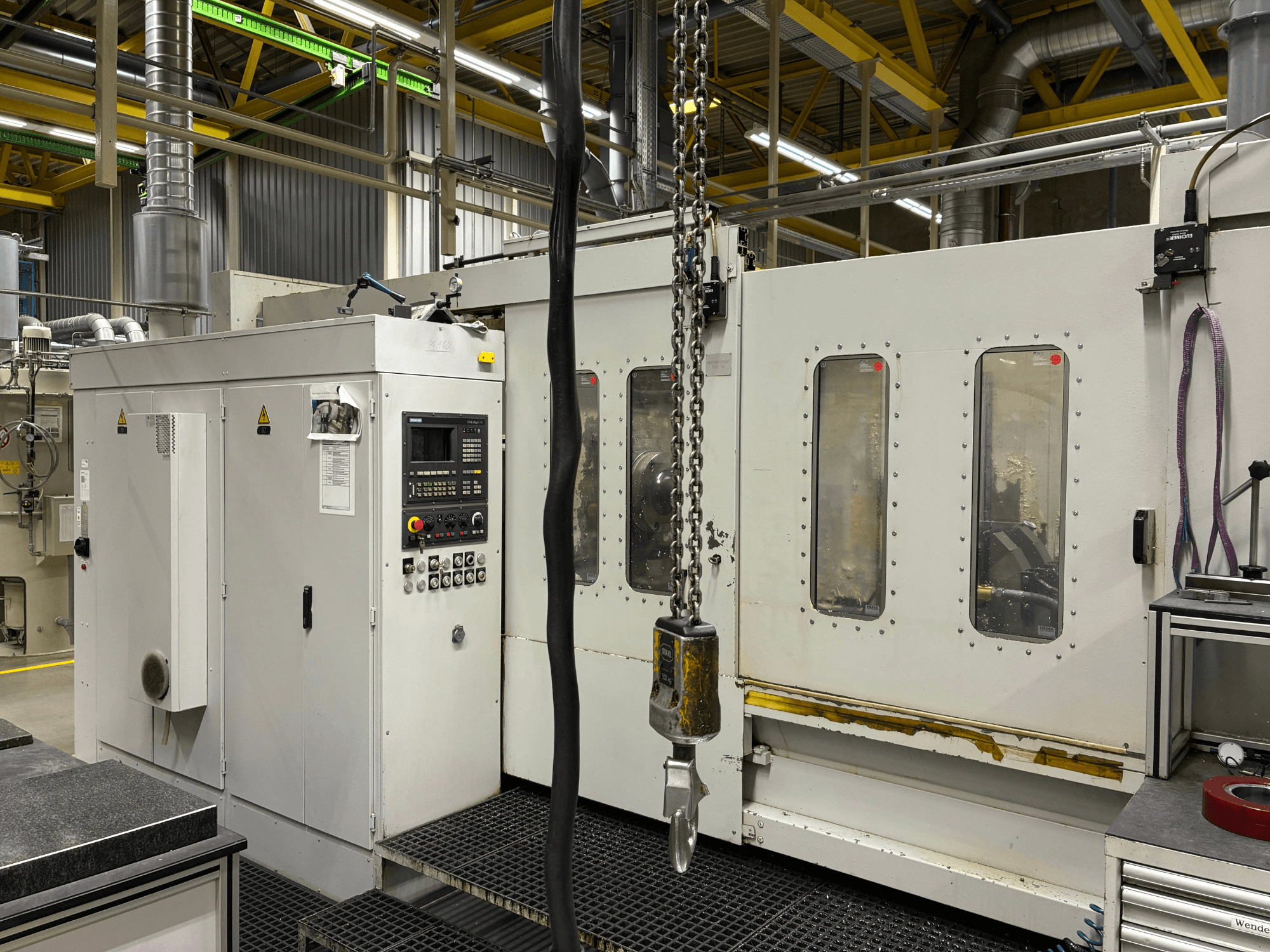 Kontrol panelli endüstriyel CNC makinesi, yandan görünüm; fabrika ortamında pencereler, zincirler ve metal bileşenler bulunur.